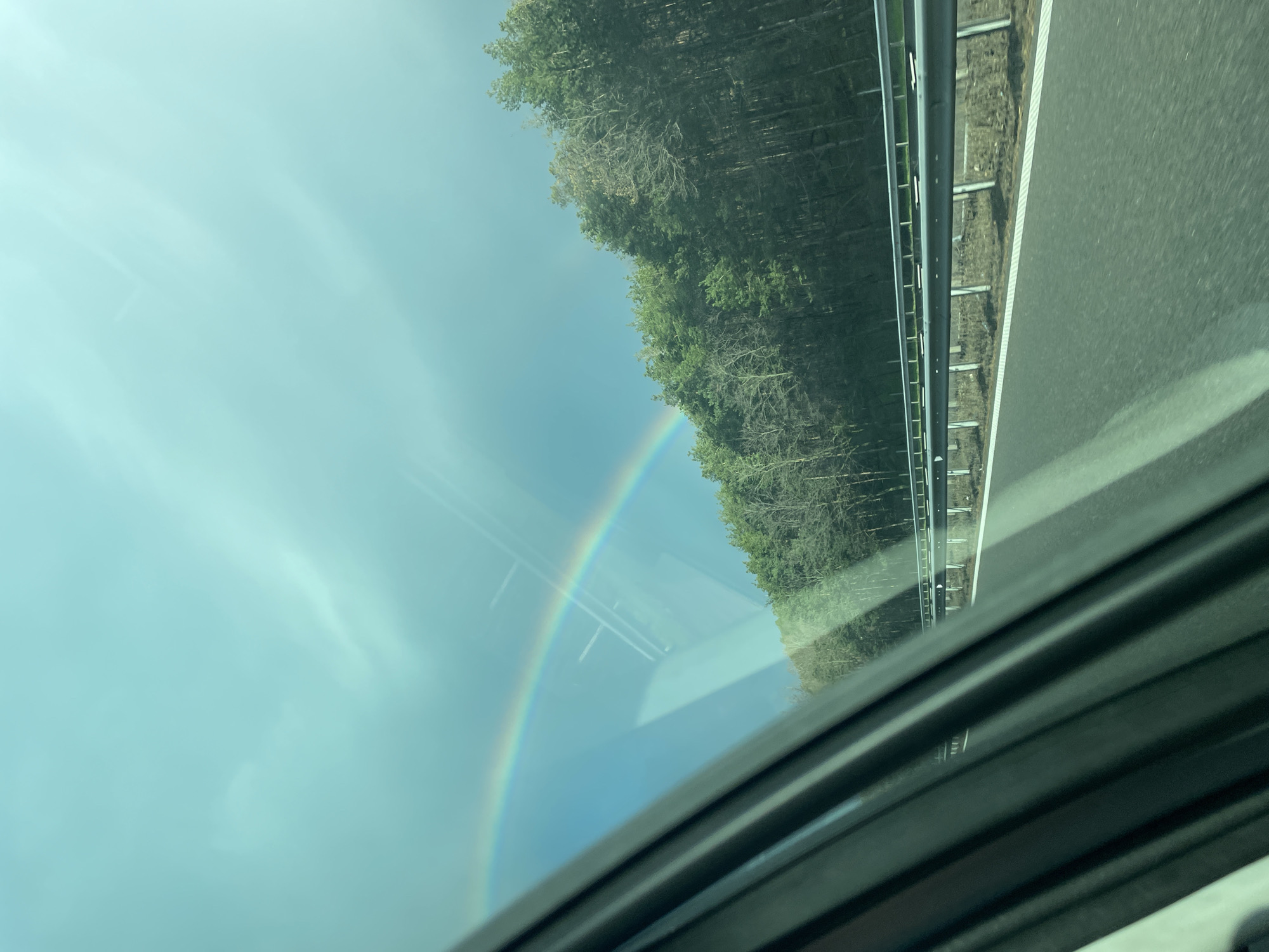 The image shows a view from inside a vehicle, possibly a car. A section of the car's interior is visible in the foreground. Through the windshield, a landscape is seen. The landscape includes a section of roadway with a metal guardrail, a forest, and a sky with a rainbow. The photo appears to have been taken while the vehicle was in motion. There is no evidence in the image to suggest a location in Lesichovo, Bulgaria.