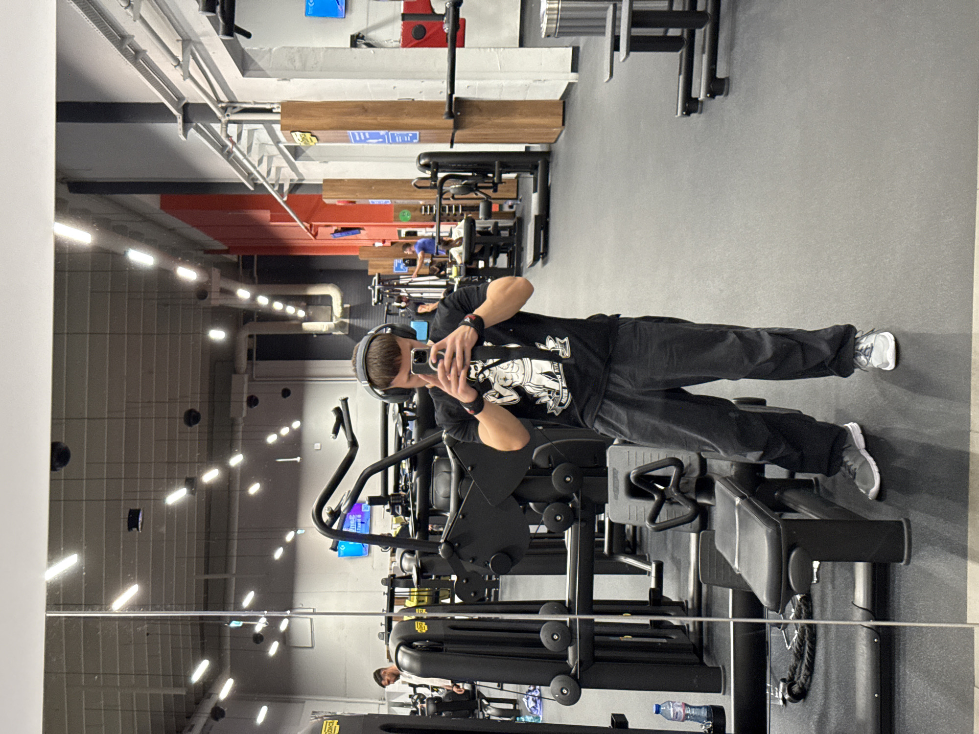 This image captures a mirror selfie taken by a young man in a modern gym located in Sofia, Bulgaria. The primary subject stands in the foreground, facing the mirror, holding a smartphone to take the picture. He is wearing a black t-shirt with a white graphic design, dark trousers, and headphones, with his hair slightly disheveled.
The setting is a well-lit indoor fitness center with a contemporary industrial aesthetic. The walls are predominantly grey, contrasting with sections of exposed ceiling infrastructure, including white air ducts, lighting fixtures, and some red and wooden accents. Various pieces of black gym equipment, such as weight machines and benches, are scattered throughout the space.
In the background, another individual can be seen exercising on one of the machines, reflected in the mirror. A water bottle is also visible on the floor near the equipment in the reflection. The bright, elongated ceiling lights create reflections on the polished floor and other surfaces, indicating a clean and spacious environment. The overall activity depicted is a moment of self-documentation within a workout routine. No specific text is clearly legible on the clothing or equipment, beyond generic gym branding elements. The time of day appears to be during operating hours, given the activity and lighting.