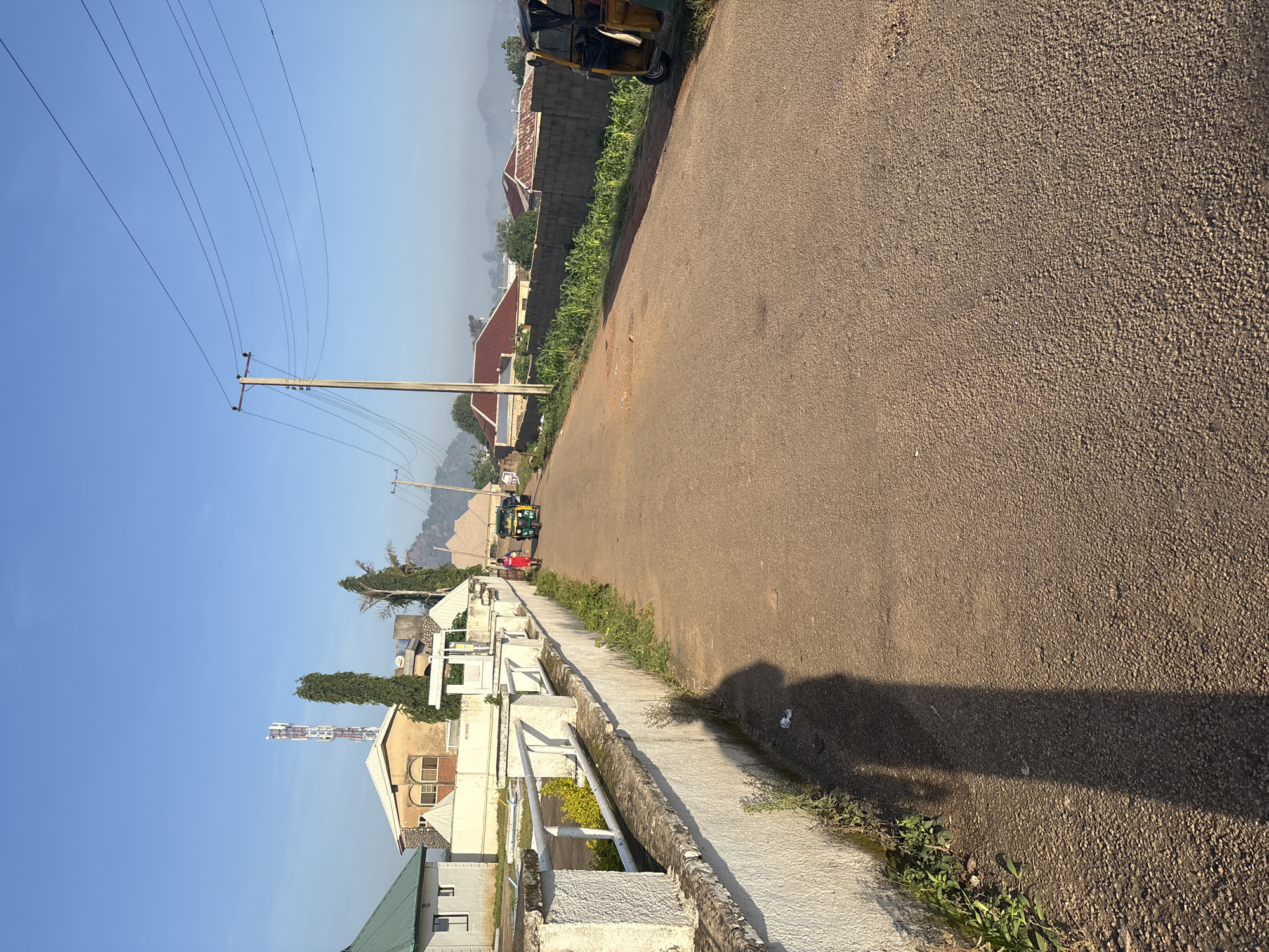 This image captures a street scene in Bukuru, Nigeria, under a clear blue sky. The road, paved with asphalt, stretches into the distance, bordered on the left by a low, white-painted wall with a metal railing. Beyond the wall are houses with various roofing materials, including green, beige, and red tiles, as well as a prominent cellular tower. On the right side of the road, a yellow, three-wheeled vehicle, commonly known as a "Keke Napep" or auto-rickshaw, is parked. Further down the road, two individuals are walking towards the parked vehicle. One person is wearing a red garment, and the other appears to be wearing a blue top and dark trousers. The foreground shows a long shadow cast by the photographer across the road and the adjacent wall. Vegetation, including grass and some leafy plants, grows along the base of the wall. The overall impression is of a residential area in a Nigerian city during daylight hours, likely in the morning given the long shadows.