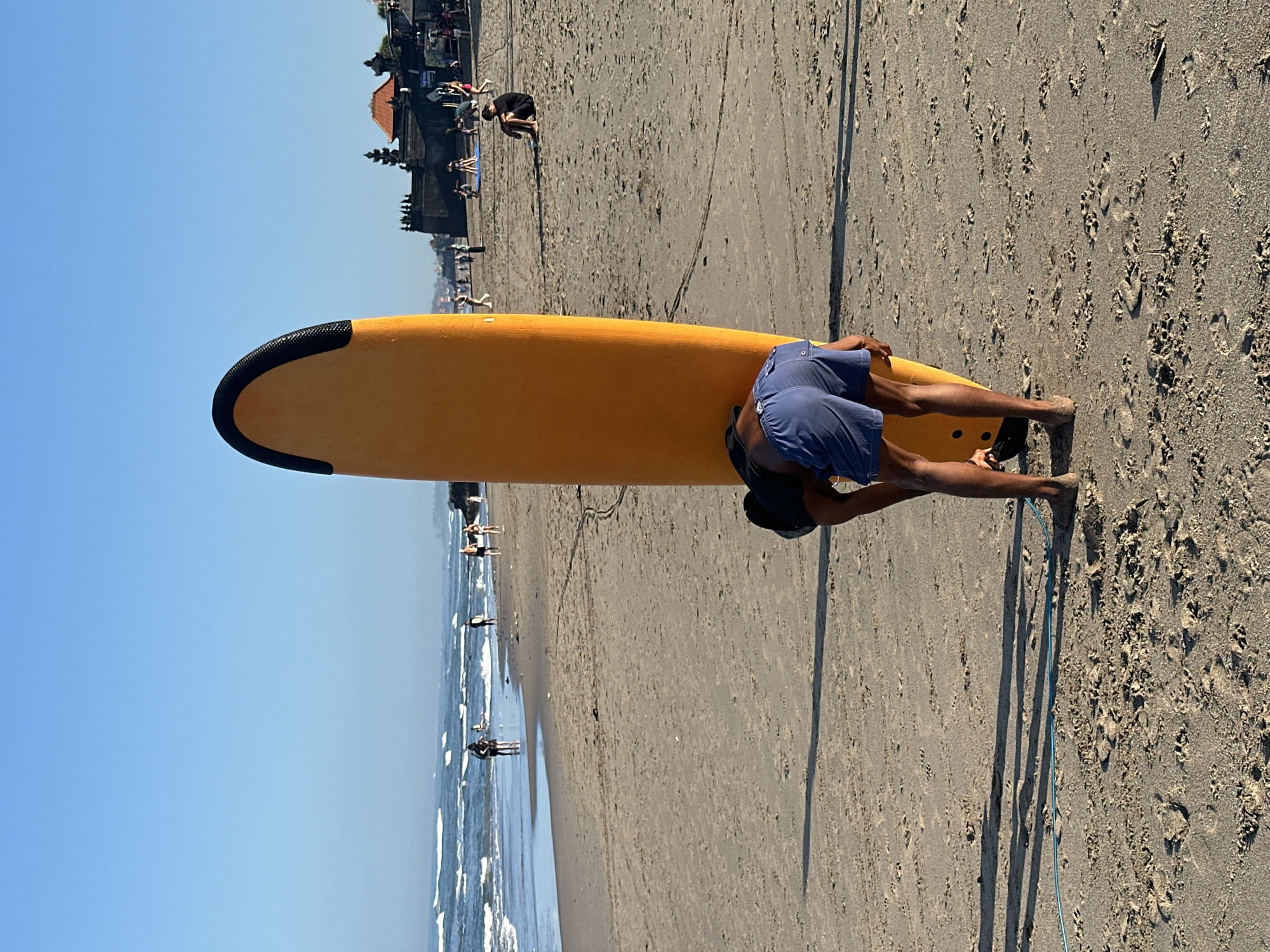 The image depicts a beach scene in North Kuta, Indonesia. In the foreground, a person wearing blue shorts bends over a tall, orange surfboard. The surfboard has a black strip at the top. The person's legs are visible, and they appear to be adjusting something on the surfboard. The beach is sandy, with some footprints and debris scattered across it. In the mid-ground, other people are visible on the beach and in the water. Some are standing, others are walking, and some appear to be swimming or surfing. The ocean has waves, and the sky is clear and blue. In the background, there are structures resembling temples or traditional Indonesian buildings. The overall scene is one of a typical beach day.