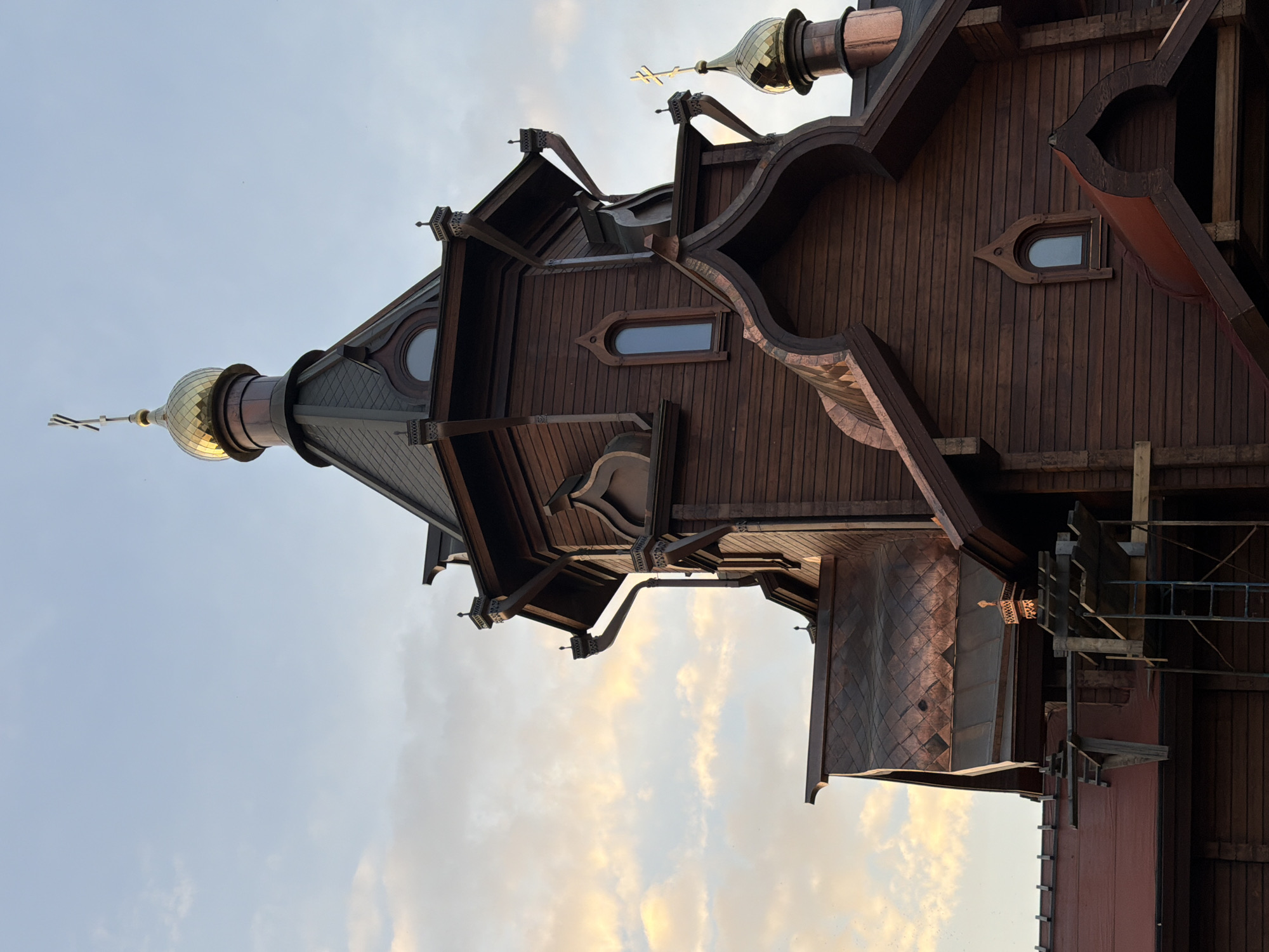 The image depicts a portion of the Church of St. Innokenty of Irkutsk in Krasnoyarsk, Russia. The church is constructed predominantly of dark brown wood, exhibiting intricate architectural details. The facade shows arched windows, layered roofs, and a golden dome with a cross. Scaffolding and construction materials are visible on the right side of the frame, indicating ongoing work or recent renovations. The background consists of a cloudy sky with a gradient of light blue transitioning to a warmer, sunset-like hue.