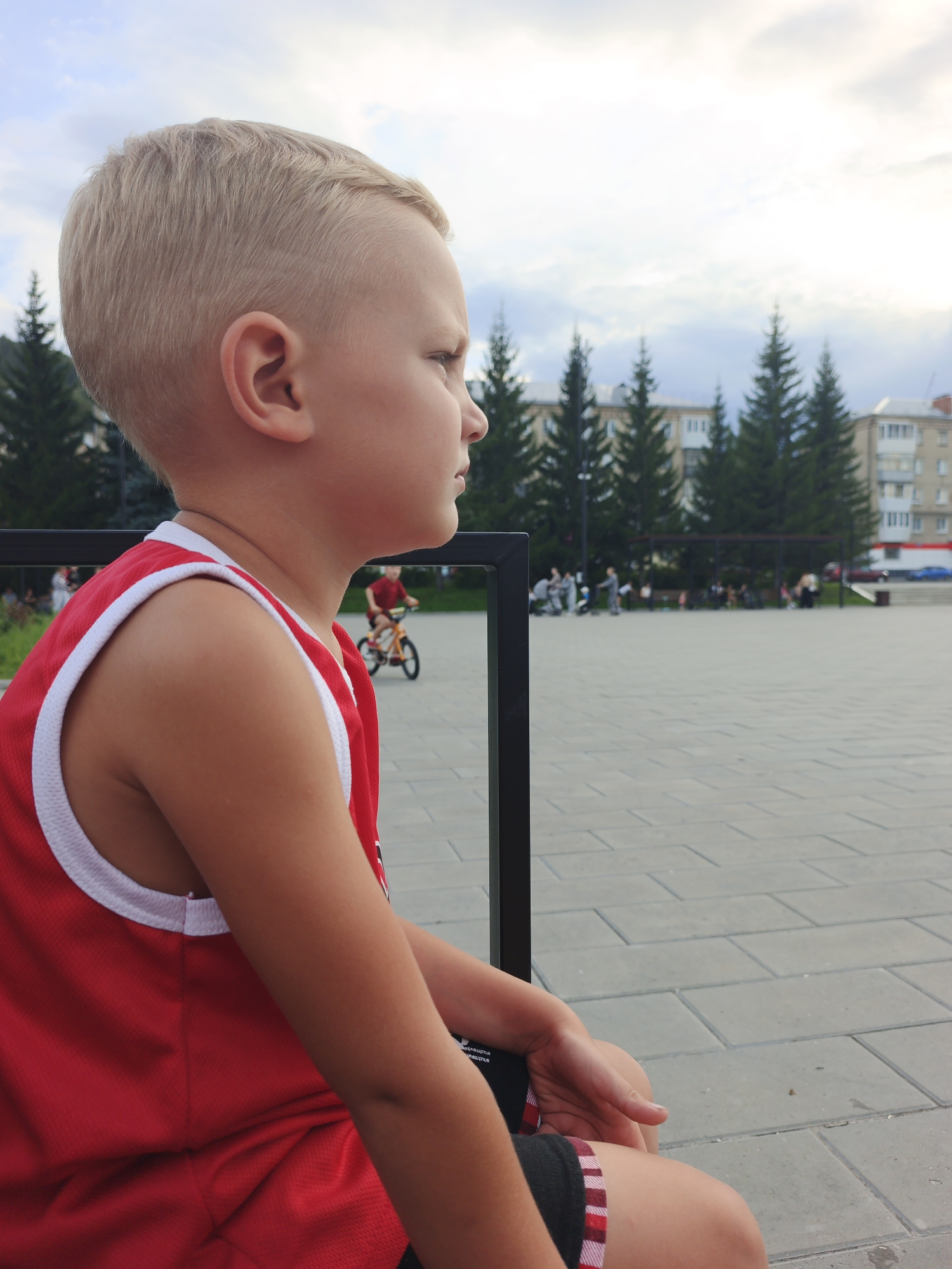 A young boy with short blonde hair, wearing a red tank top with white trim and red and black checkered shorts, sits on a bench facing right, looking out at an outdoor plaza in Zlatoust, Russia. The plaza is paved with gray interlocking stones. In the background, several tall evergreen trees stand in front of apartment buildings. Further in the background, other people are visible, including a child on a bicycle and groups of people walking. The sky is overcast with scattered clouds.