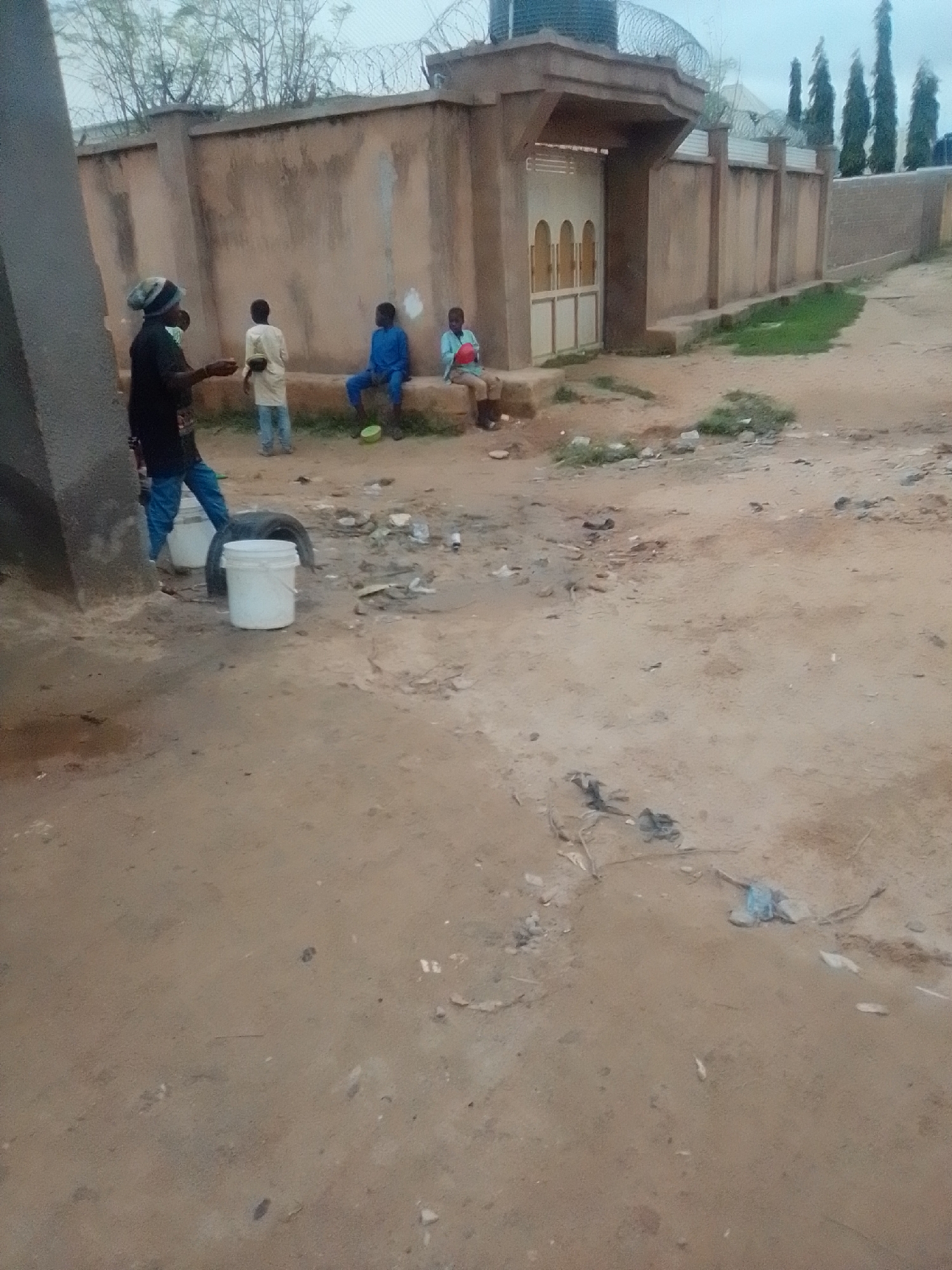 In the city of Gombe, Nigeria, a person wearing a black shirt, blue jeans, and a striped beanie is standing near two white buckets and a tire. The person appears to be interacting with someone out of frame to the left. To the right, three young boys are seated on a concrete ledge in front of a wall. One boy is wearing a blue shirt and pants, another is in a light yellow outfit, and the third is in a light blue shirt and tan pants. The environment is dusty and appears to be a residential area with a building in the background. The sky is overcast, suggesting it might be a cloudy day.