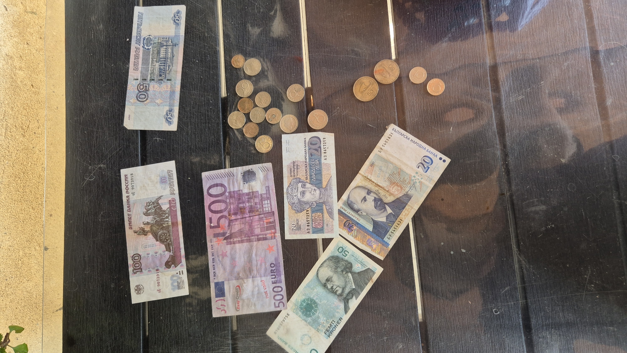 In the city of Butovo, Bulgaria, on a dark wooden table, are various currencies. A 100 Russian ruble note and a 50 Russian ruble note are placed at the top of the table. Below these are a 500 Euro note, a 50 Norwegian krone note, a 20 Bulgarian lev note, and another 20 Bulgarian lev note. Scattered around the notes are several copper-colored coins. The scene is lit by natural light, casting faint shadows.