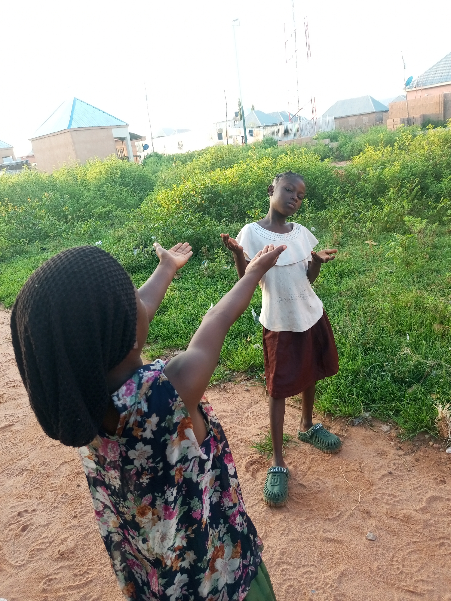 Two young girls are standing on a dirt path in a residential area of Tempure, Nigeria. The girl in the foreground, seen from behind and to the left, wears a dark, patterned dress with floral print and a black headscarf. Her arms are raised. The girl in the background, facing the camera, is wearing a white ruffled top and a brown skirt. She has her arms extended with her palms facing upwards. Behind the girls, there is overgrown green vegetation, several residential buildings with corrugated metal roofs, and a tall communication tower. The lighting suggests it is late afternoon or early morning.