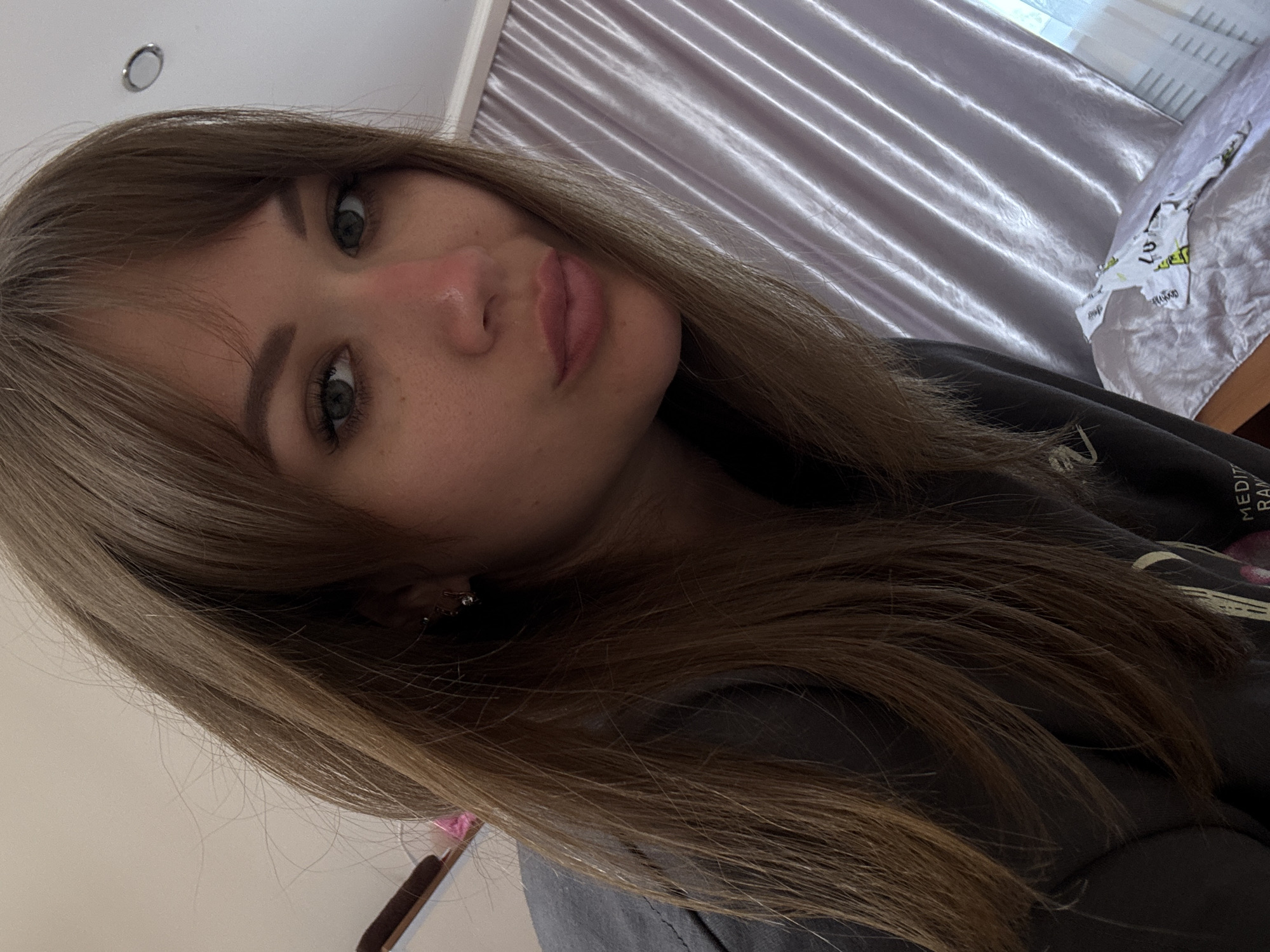 A close-up, angled selfie shows a young woman with long, light brown hair. Her hair is styled with bangs that partially cover her forehead. She has fair skin, light eyebrows, and green eyes. Her lips are slightly puckered. A small earring is visible in her left ear. She is wearing a dark grey sweatshirt. The background includes a light-colored curtain with vertical folds and a bed with white and patterned bedding. The lighting is diffused. The location, Yevpatoriya, Ukraine, cannot be determined from the visual content.
