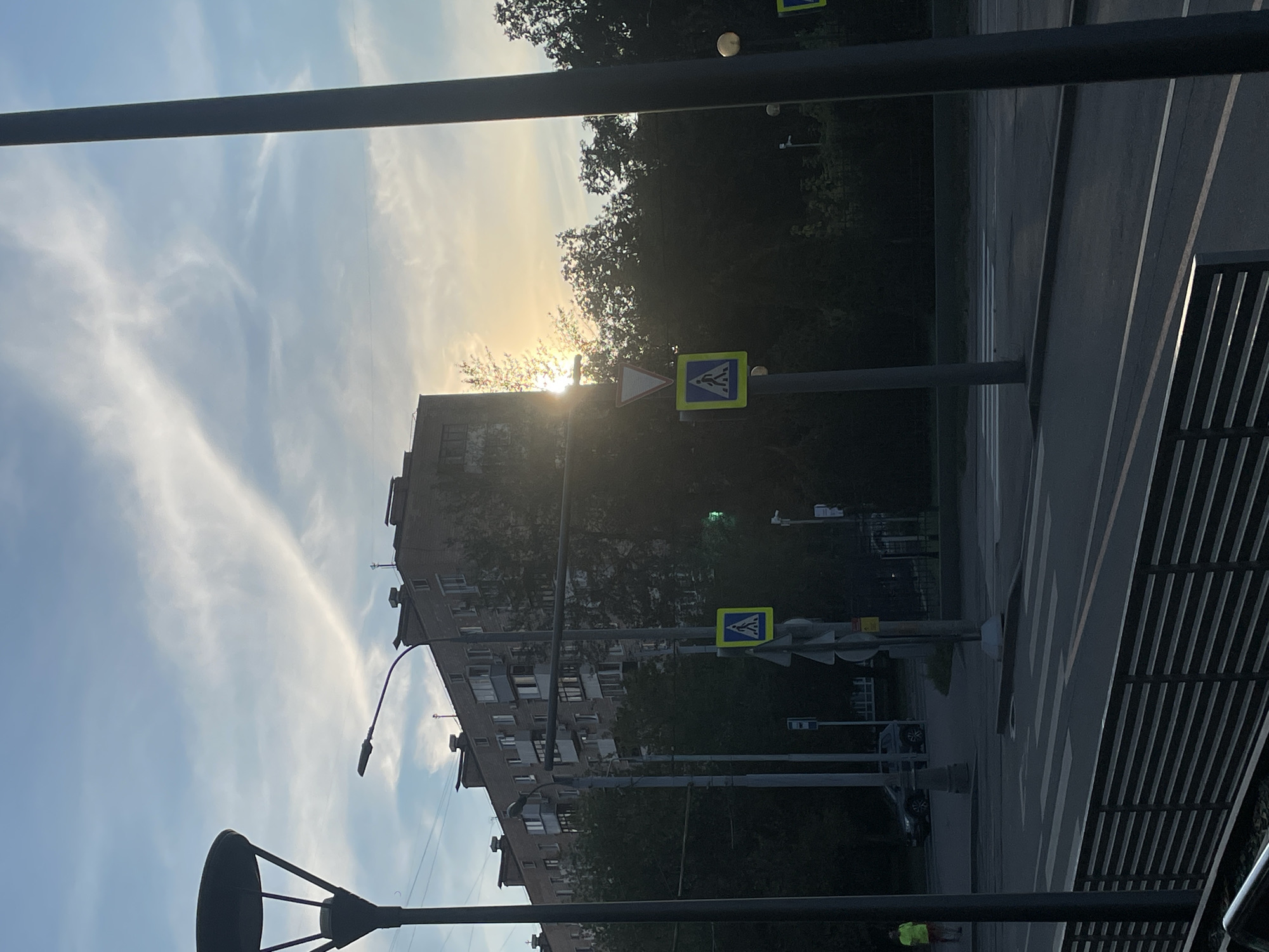 The image captures a street scene in Khimki, Russia, during the late afternoon or early evening. A multi-story brick apartment building dominates the background, with trees obscuring its lower half. Sunlight streams from behind the trees and building, creating a bright glare. In the foreground, a street with a pedestrian crossing is visible, marked by white stripes. Two pedestrian crossing signs, one yellow and square with a blue silhouette of a person walking, and another triangular red and white sign indicating caution, are positioned on poles near the crossing. A dark lamppost stands to the left, and another dark, thick pole is on the far right, obscuring part of the view. A person wearing a bright yellow shirt is standing on the sidewalk to the far left. A dark-colored SUV is parked on the street in the middle ground. The sky is a pale blue with wispy white clouds. A dark metal fence runs along the bottom of the image.