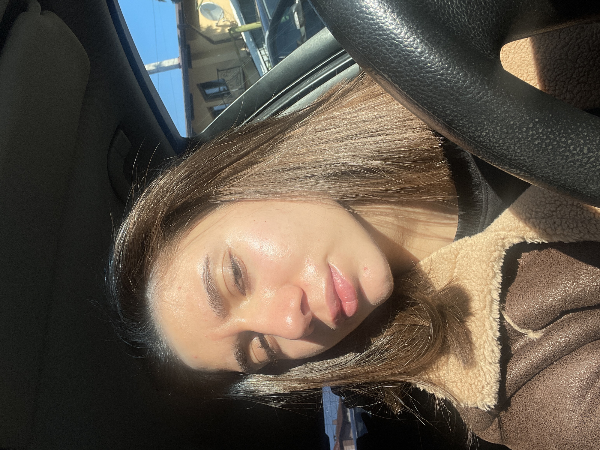 A woman is in a car. The steering wheel is visible and she is wearing a brown jacket with a beige fleece collar. The scene may be in Sofia, Bulgaria because the architecture visible through the car window is characteristic of that city.