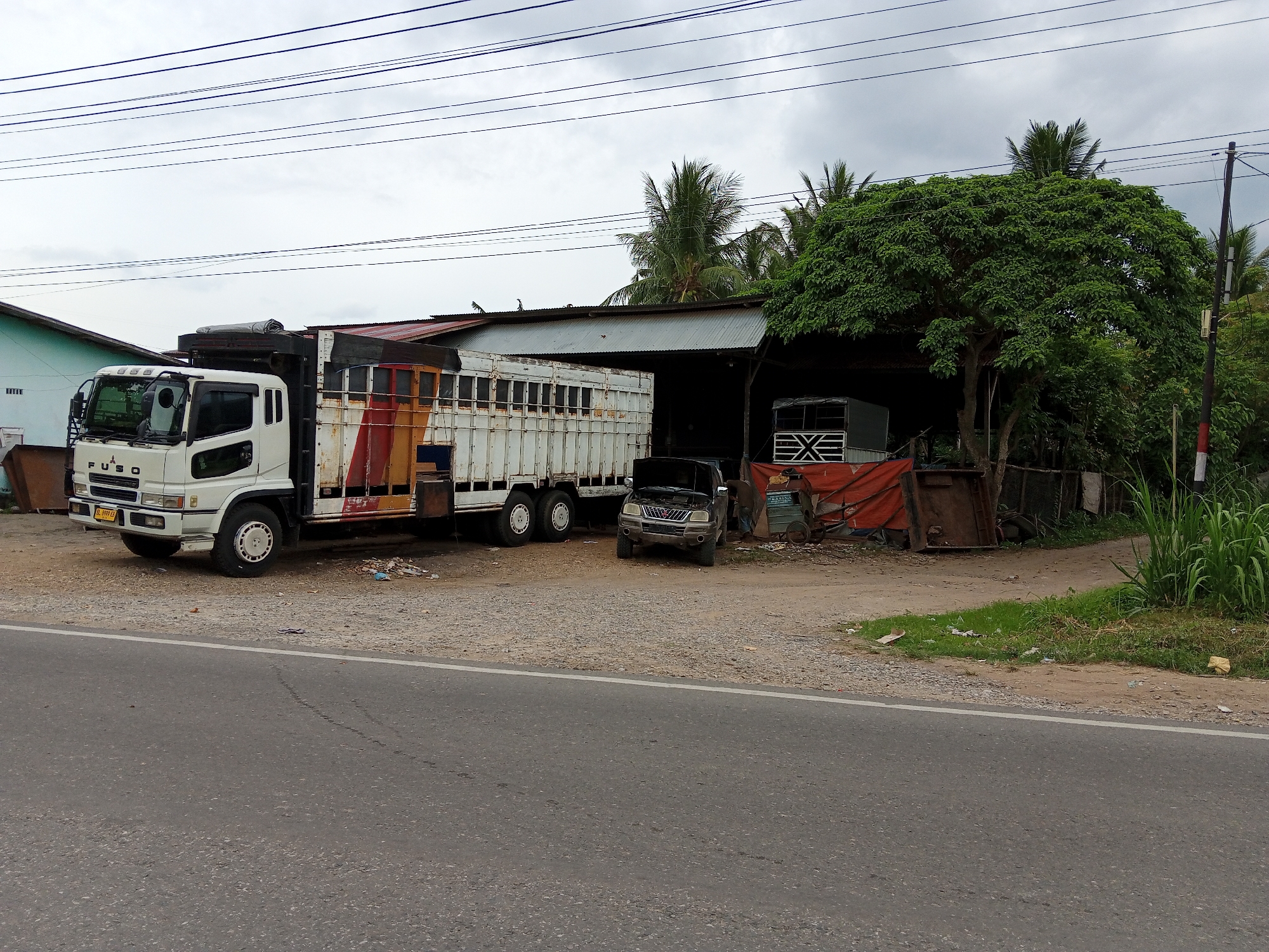 The image shows a roadside scene in Palohigeuh, Indonesia. A white Fuso truck is parked on the left, followed by a white flatbed truck with faded red and yellow markings. A dark-colored car with its hood open is parked in front of a corrugated metal structure, possibly a garage or workshop. Within the structure, another truck is partially visible. Various objects, including discarded materials and equipment, are scattered around the building and the vehicles. Palm trees and a large tree are visible in the background, along with an overcast sky. The road runs along the bottom of the image.