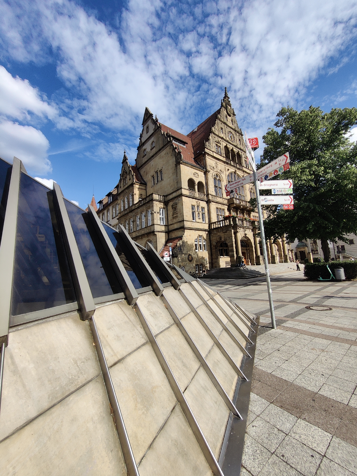 This is a low-angle, wide-shot photograph of a historic building and a modern glass structure in a public square. The building, with its ornate stone facade, multiple windows, and distinctive red-tiled roof, appears to be a town hall or similar civic structure. A grand staircase leads to the main entrance, where a few people can be seen walking and standing. In the foreground, a contemporary glass and metal structure slopes upwards, reflecting the blue sky and clouds. A signpost stands prominently in the square, adorned with various directional signs indicating distances to local landmarks such as Sparrenburg and Altstadt, as well as cycling routes. A large, leafy green tree stands to the right of the building, providing shade. The weather is sunny with scattered clouds, suggesting it is daytime. The ground is paved with interlocking stones, with a distinct pattern. A green electric scooter is parked near a trash bin. The overall atmosphere is calm and suggests a pleasant day in a European city. The visible text includes street names and route numbers on the signpost.