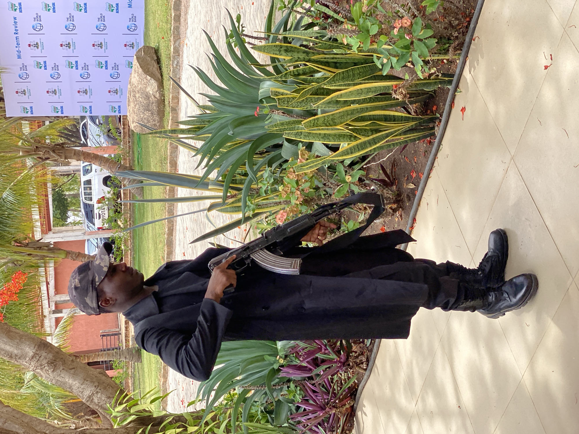 A dark-skinned man, appearing to be security personnel, stands prominently in an outdoor setting, holding an AK-style assault rifle with a silver magazine. He is dressed in a dark, possibly black, long-sleeved shirt or jacket, dark trousers, and black combat boots, topped with a camouflage-patterned baseball cap.
The scene features lush tropical vegetation, including palm trees with orange-red blossoms, agave-like plants, and variegated snake plants (Sansevieria trifasciata) with green and yellow stripes. The man stands on a light-colored tiled path, which borders a garden bed.
In the background, a large white banner is visible, indicating a formal event. Several vehicles, including a white Toyota pickup truck, are parked behind the banner. A large, natural rock is situated near the base of the banner. The bright lighting suggests it is daytime. The overall impression is an event taking place in a warm, possibly tropical, outdoor environment in Kwarau, Nigeria, with a visible security presence.
Visible text on the banner includes "Mid-Term Review," "ACRESAL," "THE WORLD BANK," and "NIGERIA" (accompanied by the Nigerian coat of arms), repeated multiple times. The word "TOYOTA" is also partially visible on one of the vehicles.