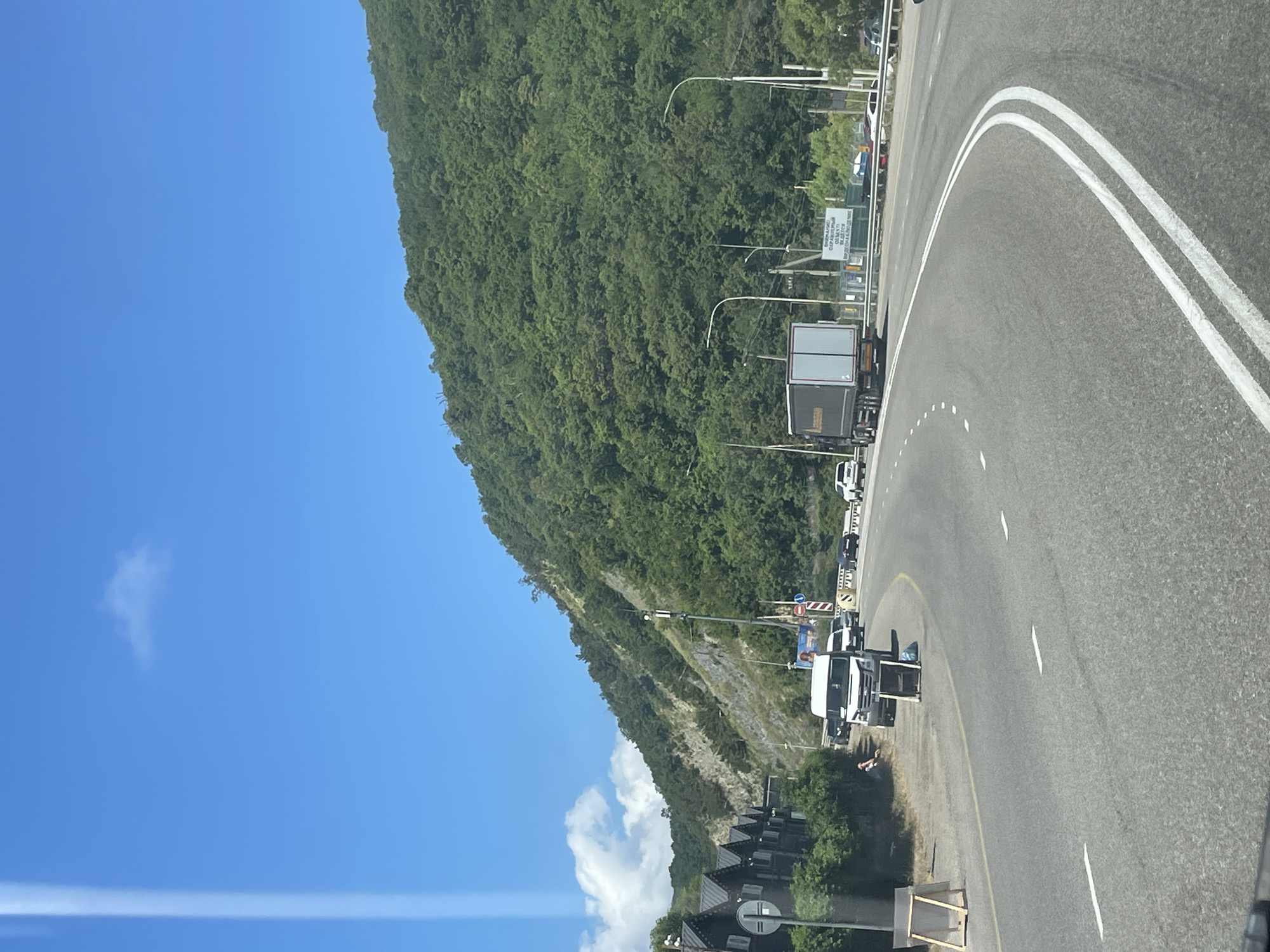 A bright, sunny day is depicted in Lermontovo, Russia, with a clear blue sky and scattered white clouds. A winding asphalt road dominates the foreground, with a truck in the right lane and several cars in the left lane. A steep, densely wooded mountain rises on the right side of the road, its green foliage contrasting with the rocky, barren slope on the left.
Along the left side of the road, a row of dark, modern houses with pointed roofs is visible. A person is sitting on the side of the road near a white van, with a wooden easel and a small table placed nearby. Road signs, including a traffic light and directional signs, are present along the roadside. The scene suggests a busy travel route through a scenic, mountainous coastal area.