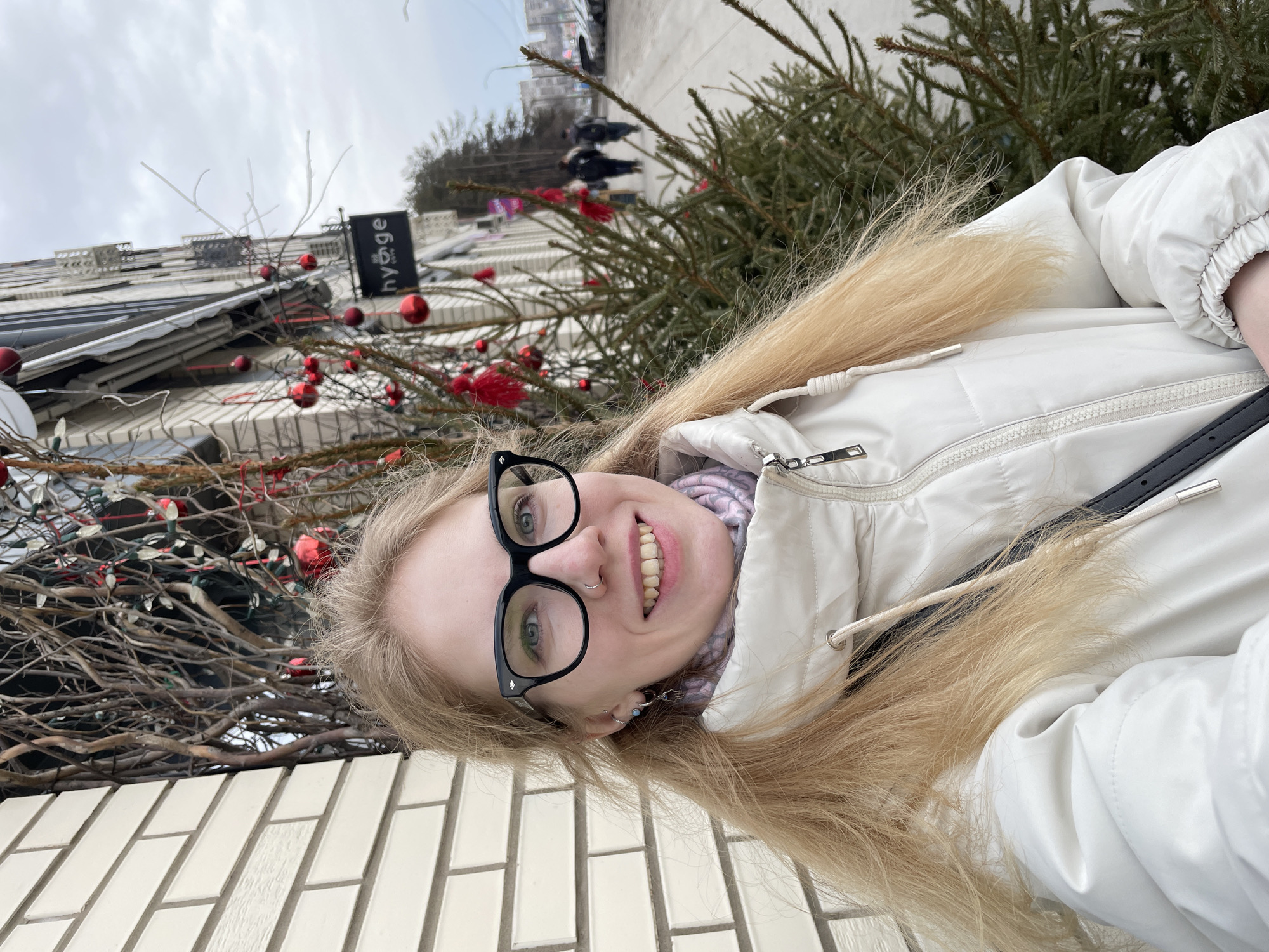 A selfie image of a young woman with blonde hair and glasses, wearing a white puffer jacket, taken outdoors in the city of Izhevsk, Russia. She is smiling. In the background, there is a building decorated with Christmas ornaments, a sign that reads "hygge", and some evergreen trees. People can be seen walking in the background. The image appears to be rotated counter-clockwise.