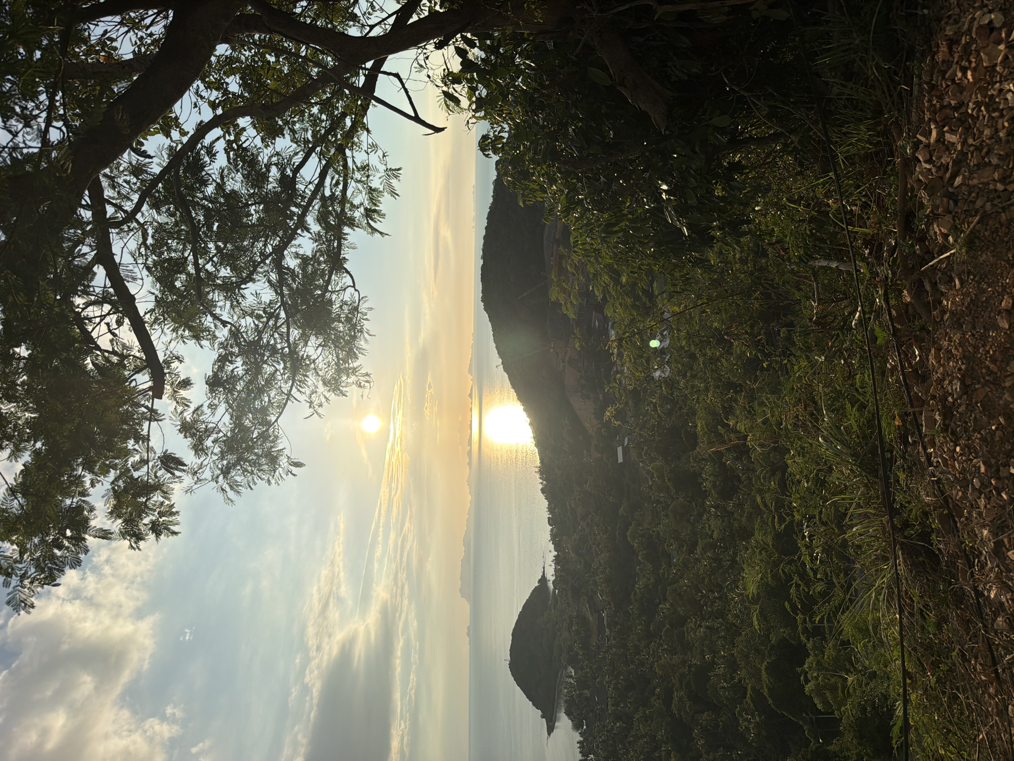 This is a vertical, eye-level, wide shot photograph of a coastal landscape at sunset. The sky is a gradient of light blue to yellow and orange hues, with wispy clouds and the sun appearing as a bright orb just above the horizon. The sun's reflection creates a shimmering path across the ocean. In the foreground, a large tree with dark branches and green leaves partially obscures the view. The middle ground features a lush, green, hilly landscape covered in dense vegetation and some scattered buildings. A small island with a pointed hill and a structure on top is visible in the ocean to the left of the frame. In the distance, a chain of islands can be seen across the water. The ground in the immediate foreground is rocky and uneven, with dry dirt and small stones. The scene is set in Baan Thong Po, Thailand, a location characterized by its tropical coastline and abundant greenery. There are no people or animals visible in the image. The overall atmosphere is serene and natural, capturing the beauty of a tropical sunset.