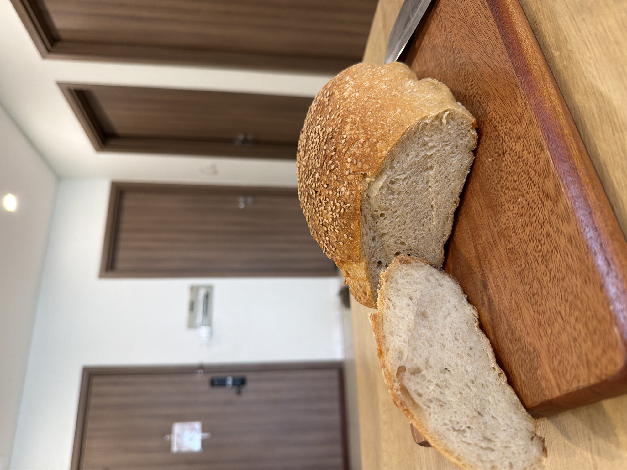 An indoor, close-up, and slightly overhead shot captures a rustic loaf of bread, partially sliced, resting on a wooden cutting board. The bread features a golden-brown crust liberally coated with sesame seeds, revealing a light, airy crumb with an irregular texture from the two visible slices. One slice is fully detached, lying next to the main loaf on the board. A silver knife handle is partially visible at the top edge of the cutting board, suggesting recent slicing.
The cutting board itself is made of a dark, reddish-brown wood with a clear grain, placed on a lighter wooden or laminate surface. The background is a brightly lit interior space, possibly a hallway or room in a residence or hotel in Bac Nha Trang Ward, Vietnam. Multiple closed, dark brown wooden doors with recessed panels are visible against white walls. A small, blurry rectangular object is mounted on the wall near one door, and what appears to be an indistinct, light-colored sign or sticker is on another door. The overall impression is one of a clean, modern, and functional indoor setting. No legible text is visible.