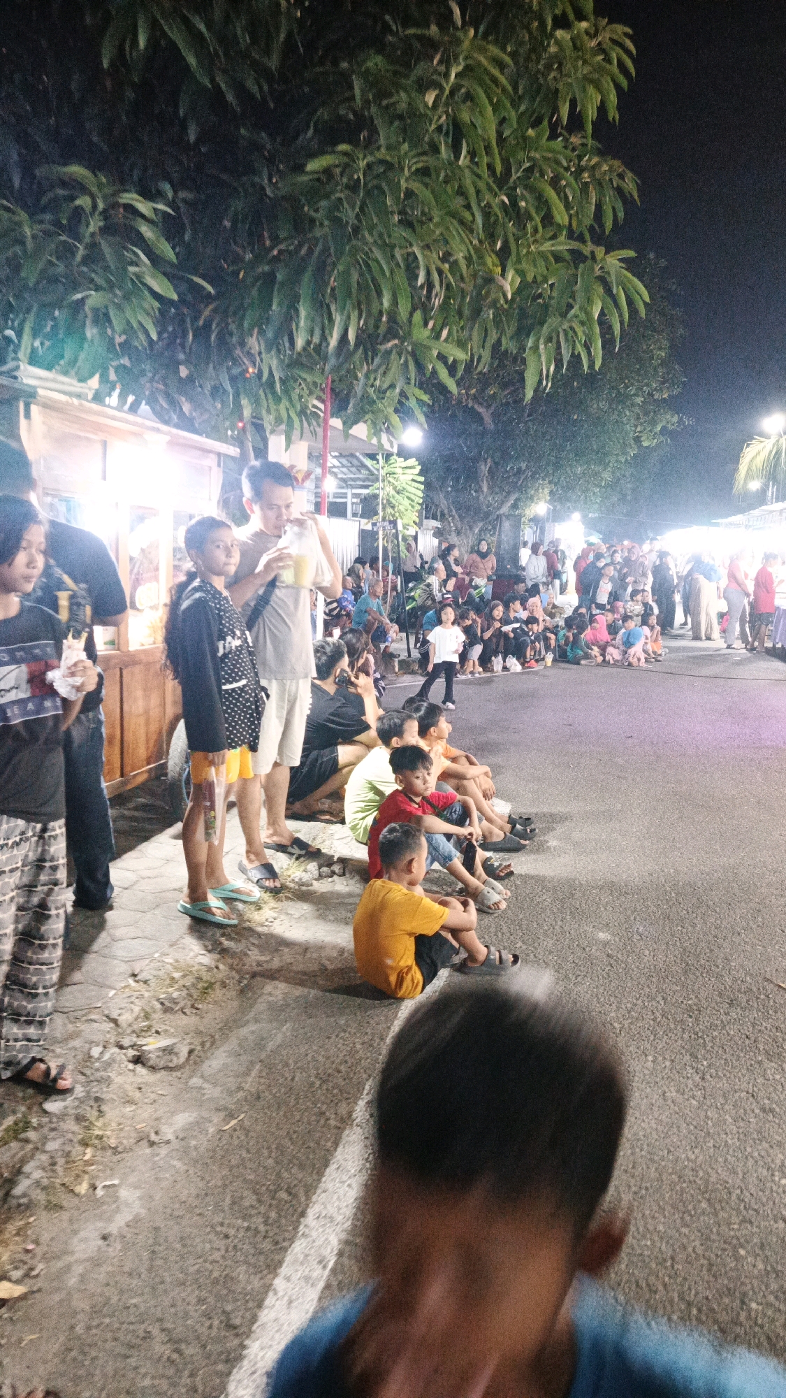 The image depicts a nighttime scene in the city of Surakarta, Indonesia, likely a public gathering or street event. A group of people, including adults and children, are gathered along the side of a road, facing away from the viewer. Many are seated on the curb or directly on the asphalt, some with their backs to the camera. The foreground shows the blurred back of a child's head, obscuring their face.
To the left, under the canopy of large, leafy trees, a food stall is visible with bright lighting. Several individuals are standing near the stall, including a man holding a clear plastic bag filled with a light-colored liquid, possibly a drink. A young girl in a black and white patterned shirt stands next to him. Further left, a girl in a black t-shirt and patterned pants holds a white object in her hand.
The people seated on the curb are diverse in age, with several young boys prominently in the foreground wearing casual clothing like t-shirts and shorts. The background shows more people gathered, some appearing to be watching something happening further down the street, indicated by the direction they are facing. Lights from stalls and streetlights illuminate the scene, creating a festive atmosphere. The presence of trees and the style of the stalls suggest a tropical urban environment.