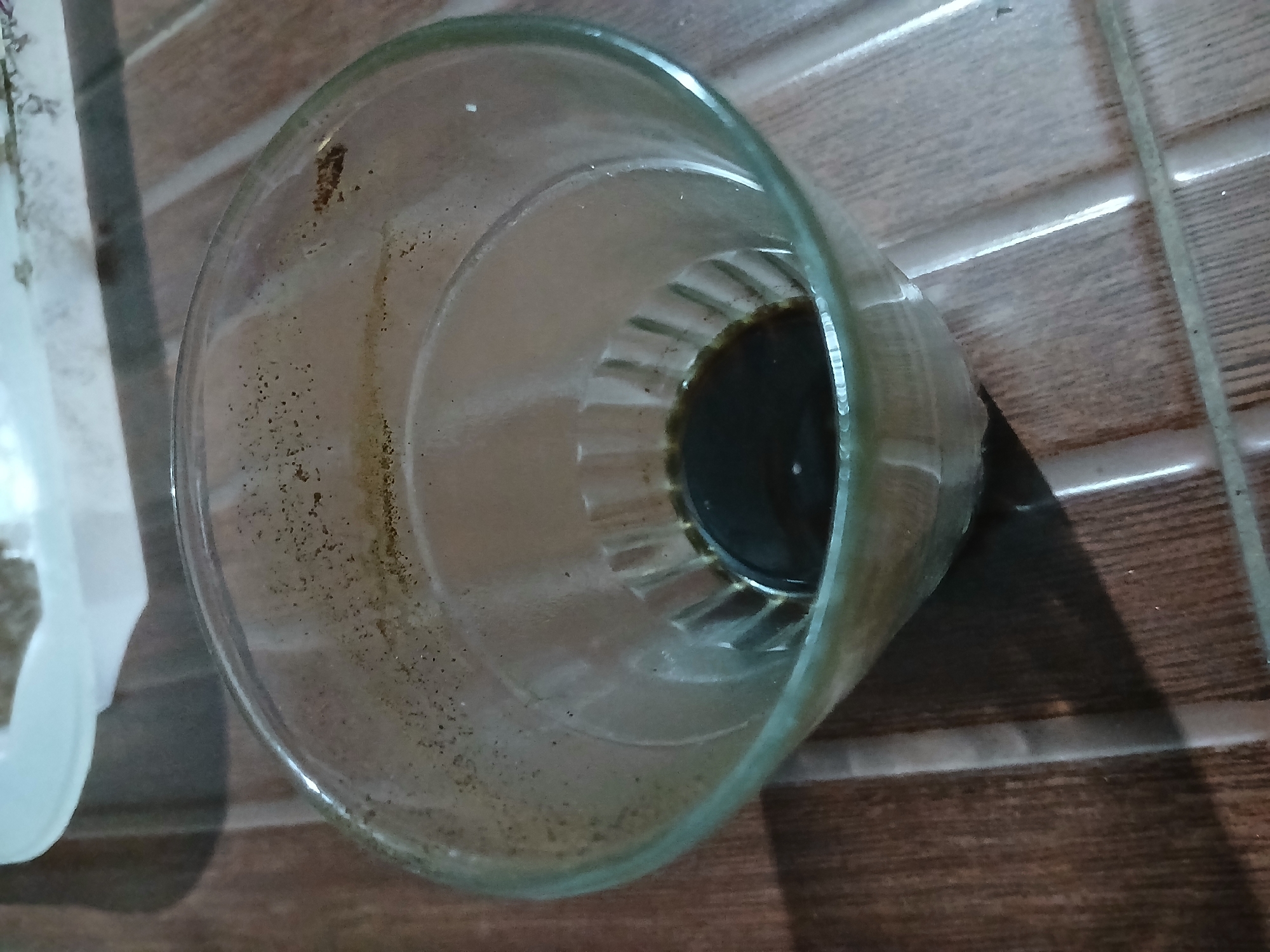 A close-up shot shows a glass with a small amount of dark liquid at the bottom, possibly coffee residue. The glass is cylindrical with a ridged pattern on the lower interior. The exterior of the glass has some small specks of dirt or residue. The glass sits on a tiled surface with a wood-like pattern. In the upper background, a white object, possibly a plate or lid, is partially visible. The scene is well-lit, suggesting it was taken indoors. The location is Sekarputih, Indonesia.