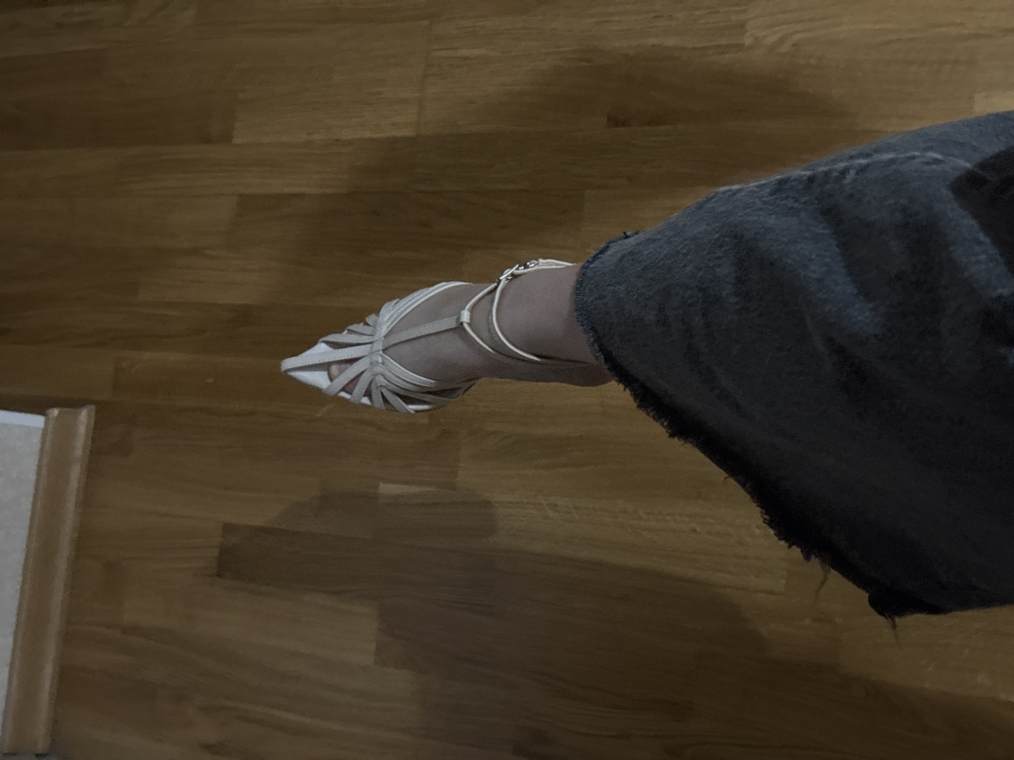 This media file captures a close-up, angled view of a person's lower leg and foot, wearing a stylish shoe on a wooden floor. The foot is adorned with a light-colored (possibly white or cream) strappy high-heeled sandal, featuring multiple thin straps crisscrossing over the toes and instep, leading to an ankle strap. A small, silver-toned metallic clasp or decorative element is visible on the outer side of the ankle strap. The lower part of the leg is clad in a dark-colored garment, likely denim jeans, characterized by a slightly frayed or distressed hemline.
The setting is an indoor space with a polished wooden floor, displaying distinct planks and wood grain. The lighting appears somewhat muted, casting subtle shadows on the floor around the foot and leg. The pose suggests the person is extending their leg forward, possibly to showcase the footwear or in the midst of a natural movement. No specific activity beyond this is clearly defined. There is no visible text in the image. The scene is situated in Chaikovsky, Russia, as provided in the context.