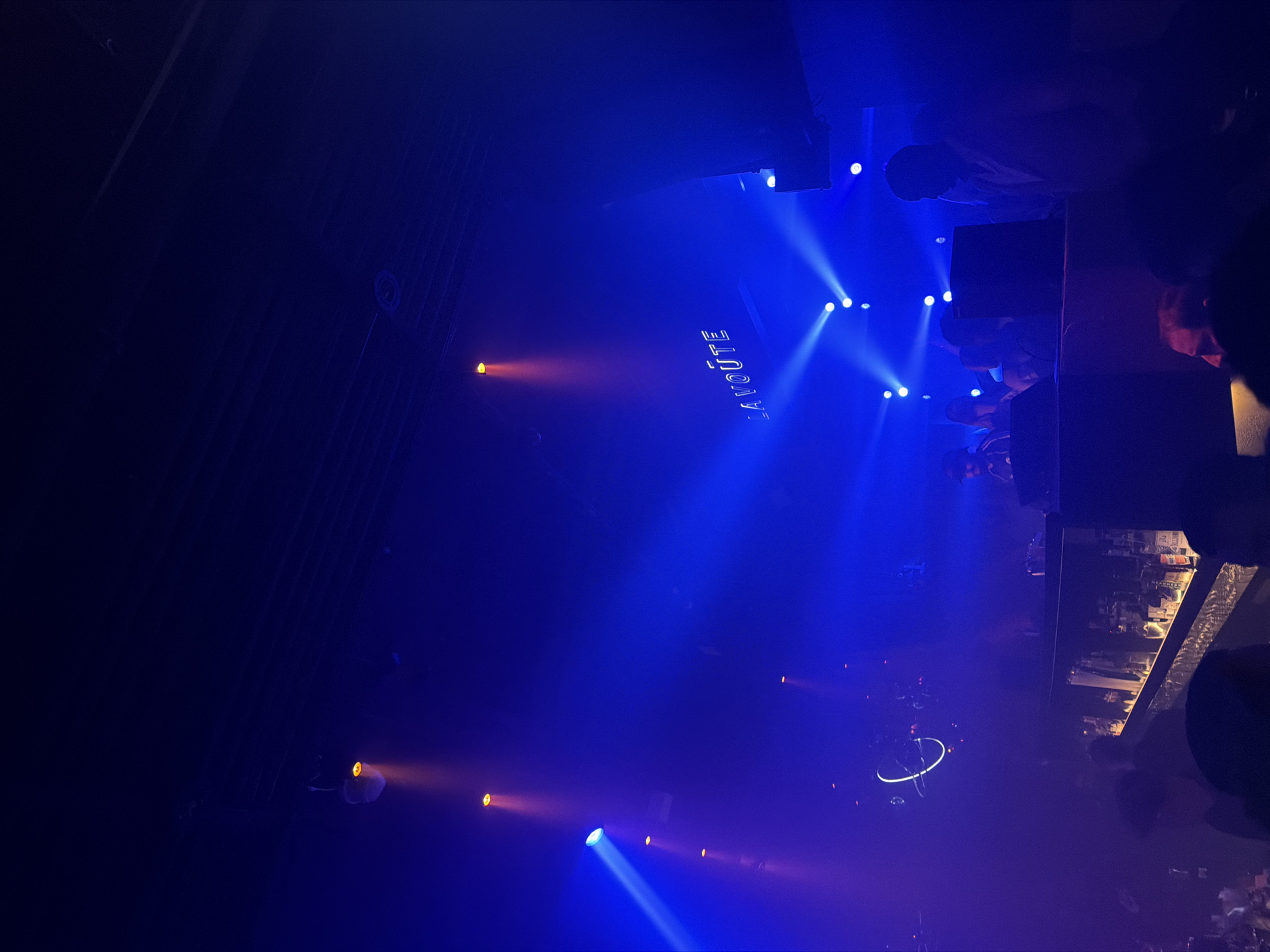 The image depicts a dimly lit nightclub scene, likely La Voûte, located in Montreal, Canada, evidenced by the illuminated text "LA VOÛTE" on a dark surface. The atmosphere is dominated by vibrant blue and purple lighting, with beams of light cutting through the smoky air, illuminating a dance floor area. In the foreground, a bar with bottles and glassware is visible, suggesting a social gathering or event. Several individuals are present, some silhouetted against the light, suggesting they are patrons or staff. One person appears to be standing behind the bar, possibly tending to drinks. Another individual, silhouetted and wearing a suit, stands near the bar. In the mid-ground, a DJ setup with speakers and equipment is present, with a figure behind it, indicating a musical performance. The ceiling is dark and industrial, with visible light fixtures casting beams downwards. The overall impression is that of a lively nightlife establishment.