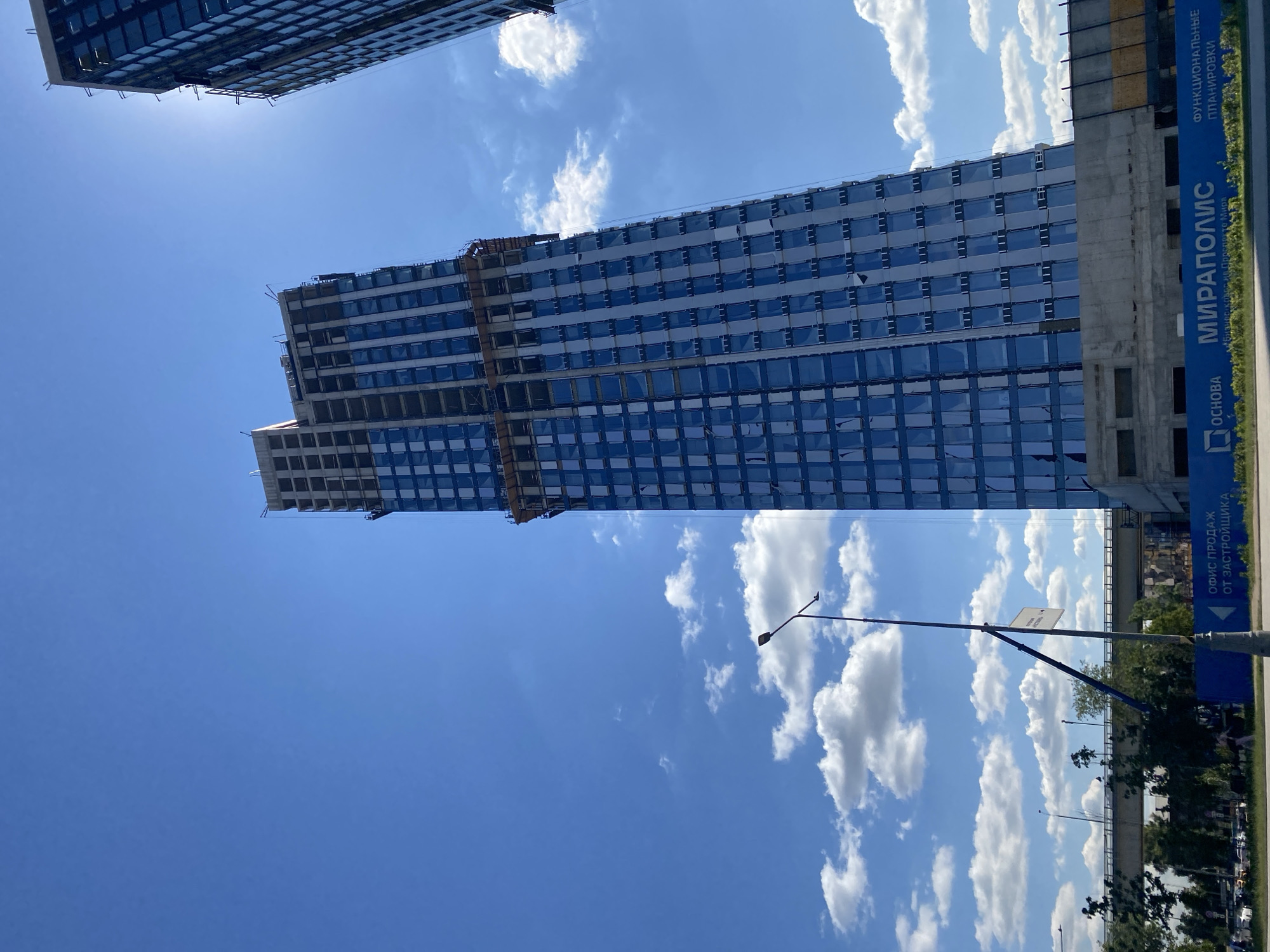 An outdoor, low-angle, vertical medium shot captures a skyscraper under construction against a bright blue sky with scattered white clouds. The skyscraper, clad in blue glass panels, is the primary subject. Construction scaffolding is visible around the upper floors. To the right, another partially visible skyscraper under construction is seen, with the sun glinting off its glass facade. In the foreground, a lamppost stands to the left of the main skyscraper. A blue construction barrier runs along the bottom of the frame, displaying text in a non-English script. Beyond the barrier, a concrete overpass or bridge is visible, with trees and vegetation lining the area below it. The overall scene suggests an urban development area in the city of Unknown.