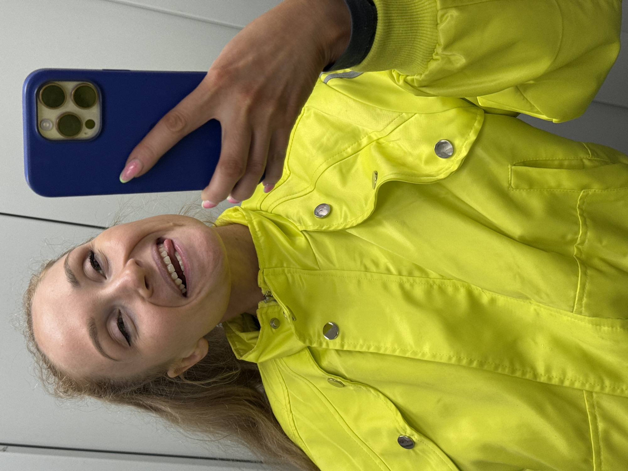 A fair-skinned woman with light brown hair is taking a selfie with a dark blue smartphone, presumably in front of a mirror. She's wearing a bright yellow windbreaker-style jacket with metallic button closures and a zippered front. The camera of the phone has four lenses. The location cannot be identified.
