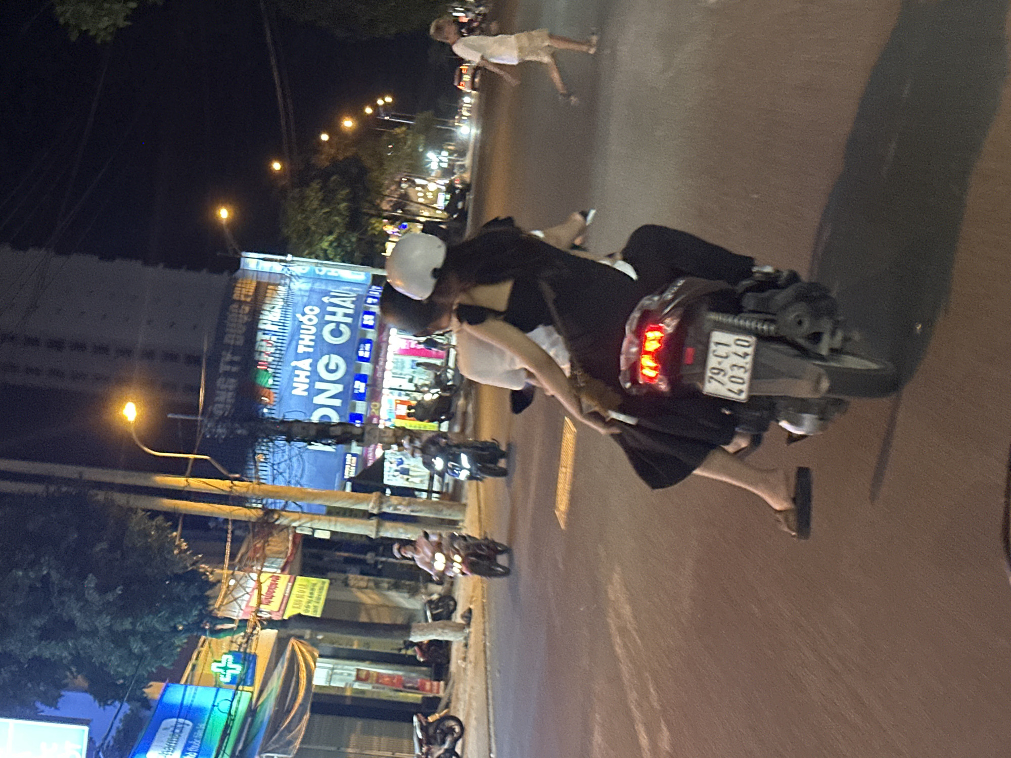 The image shows a street scene at night in Bac Nha Trang Ward, Vietnam. A motorcycle with two people is prominent in the foreground. The motorcycle's license plate reads "79-C1 40340". The driver is wearing a white helmet and dark clothing, while the passenger wears light-colored clothing. The passenger is holding onto the driver. Both individuals are wearing sandals. Other motorcycles are parked along the side of the road, and a person is walking on the sidewalk in the distance. Buildings with lit signs, including one for a pharmacy ("NHA THUOC DONG CHAU"), line the street. A streetlamp and power lines are also visible.