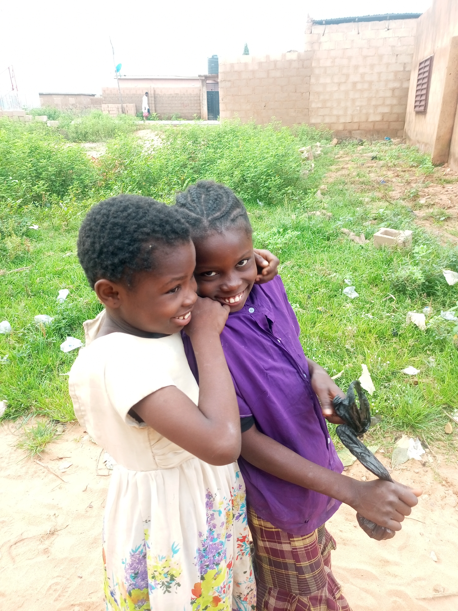 Two young Black girls are posing for a photograph outdoors, with one girl embracing the other. The girl on the left is wearing a light-colored dress with a floral pattern, and the girl on the right is wearing a purple button-up shirt and a plaid skirt. The background appears to be a rural or undeveloped area, possibly on the outskirts of Tempure, Nigeria. There is overgrown green vegetation and some sandy ground. In the distance, there are buildings constructed with brick and concrete, characteristic of Nigerian architecture, and a water tank is visible on top of one of the structures. The sky is overcast.
