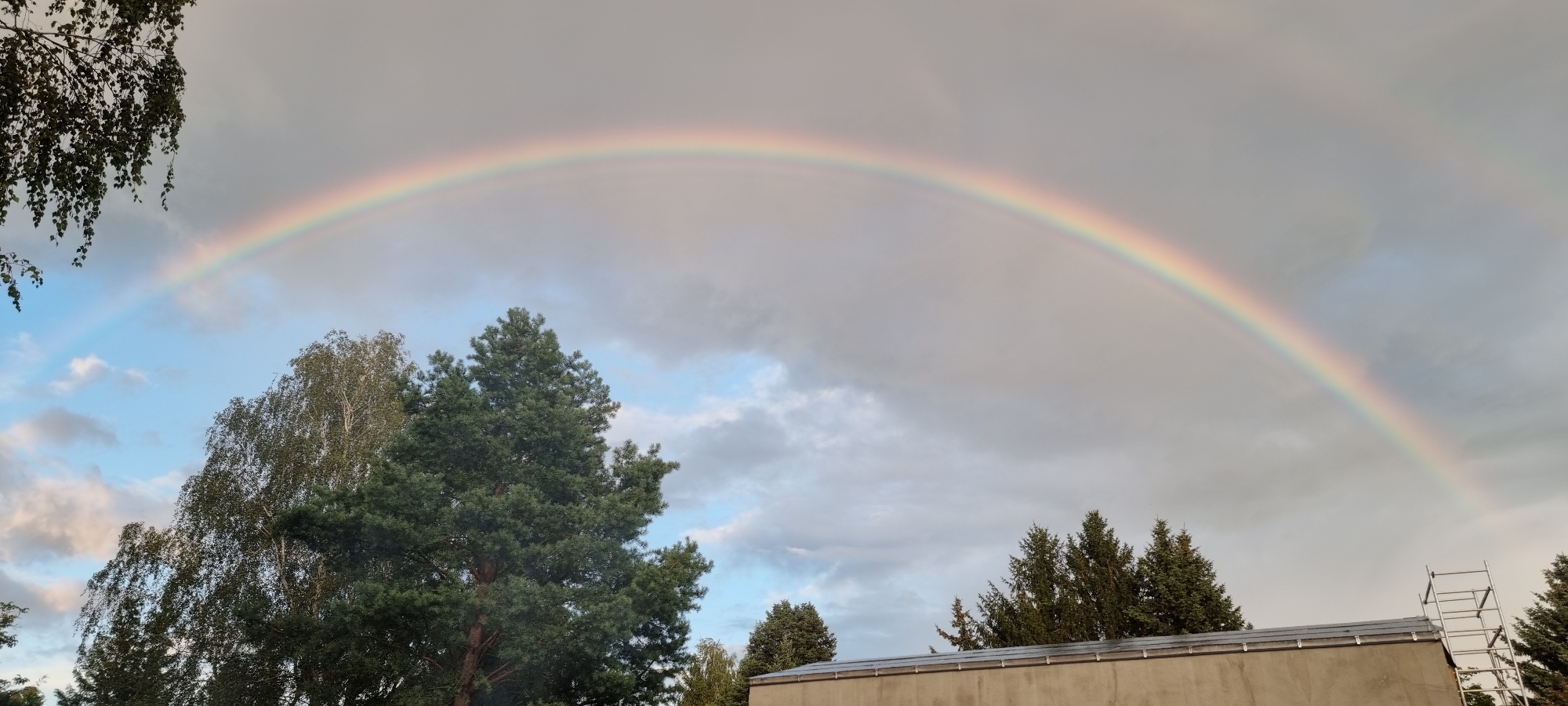 A vibrant rainbow arcs across a cloudy sky over Szczaki, Poland. The sky is a mix of grey clouds and patches of light blue, with the rainbow displaying its full spectrum of colors. In the foreground, several trees are visible, including a large pine tree with dark green needles and a deciduous tree with lighter green foliage. To the right, the top of a building with a grey roof and a section of a metal scaffolding structure are present. The scene captures a moment of natural beauty following rain.