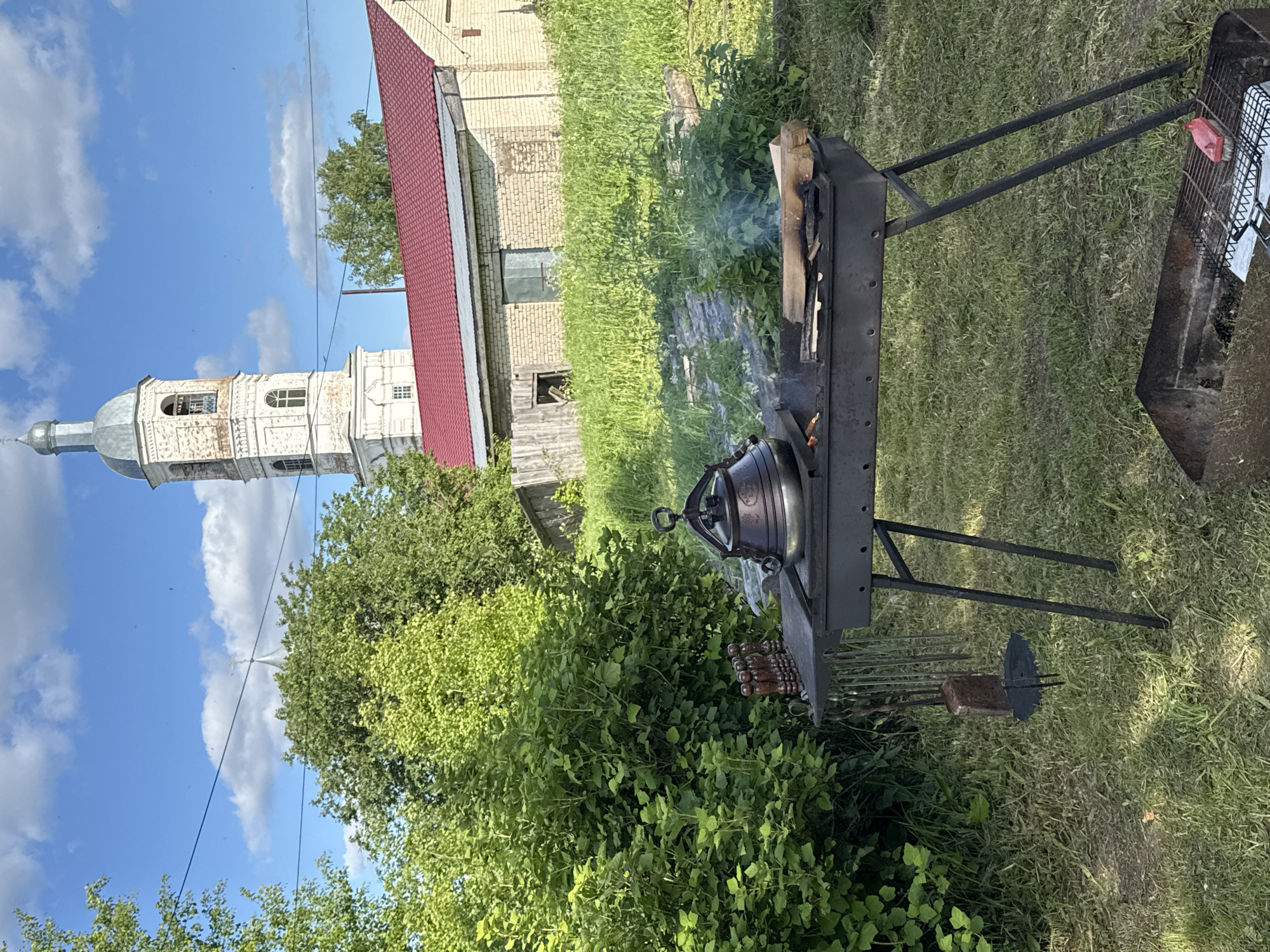 The image captures an outdoor scene set in what appears to be a rural or semi-rural environment in Луптюг, Russia, under a clear blue sky with scattered white clouds. The ground is covered with green grass and lush bushes, suggesting a warm, sunny day.
In the mid-ground and foreground, an outdoor cooking setup is visible. A dark metal brazier or grill stand holds a cast-iron kettle, possibly a kazan or Dutch oven, placed directly over burning wood and coals. Smoke gently rises from the fire, indicating active cooking. A large piece of wood is still burning on the left side of the brazier. Below the kettle, several metal skewers with wooden handles are arranged in a holder, suggesting preparations for grilling meat or vegetables. To the right of the kettle, another section of the grill features a metal grate, on which a red object (possibly a piece of food or a container) rests next to a white cloth or paper. Further right on the ground, a metal tool, resembling a shovel or ash rake, is present.
In the background, a striking architectural element dominates the left side of the frame: a historic, light-colored bell tower or church steeple with a domed, silvery roof. Its weathered facade suggests age and historical significance. Behind the bell tower and extending towards the right, a building with a bright red gabled roof and light-colored brick walls is partially visible. The overall scene depicts a peaceful moment of outdoor cooking, likely a leisure activity, with a backdrop of traditional Russian architecture and nature. There is no visible text in the image.