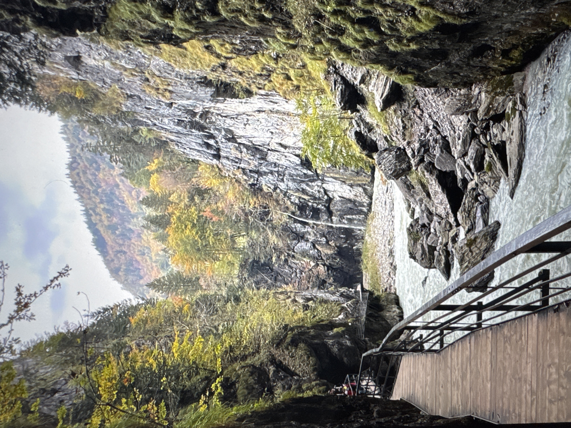 The image depicts a scenic gorge with a river flowing through it. A wooden walkway with a metal railing runs alongside the gorge, offering a path for visitors. The rocky walls of the gorge are covered in moss and vegetation, with trees displaying autumn colors on the slopes above. The river appears to be fast-flowing, with rocks and boulders scattered in its path. The sky is overcast. The scene appears to be a natural landscape, possibly a tourist attraction for hiking or sightseeing. There are no specific landmarks or features that would indicate it is located in the city of Porrentruy, Switzerland.