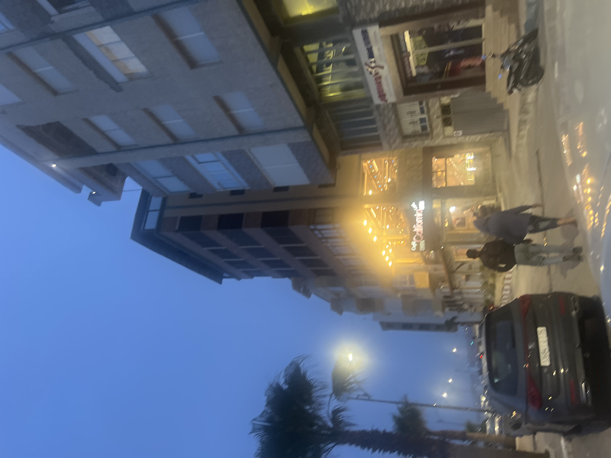 The image depicts a street scene in Fnideq, Morocco, taken during the evening. Tall buildings line the street, some with visible storefronts. Pedestrians are walking on the sidewalk. A parked car is visible in the foreground. The sky is a dark blue, suggesting it is either dusk or dawn. Palm trees are visible in the lower part of the frame, adding to the locale. Streetlights illuminate the scene.