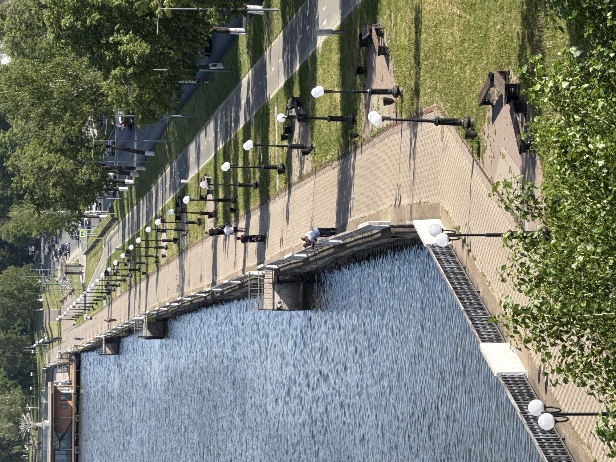 The image depicts a waterfront scene in Votkinsk, Russia. A paved promenade runs along the water's edge, lined with evenly spaced lampposts. Adjacent to the promenade is a walkway and grassy area with benches under trees. The water surface is rippled, reflecting the sky. Several people are visible on the promenade, appearing as pedestrians. The image is taken from an elevated perspective.