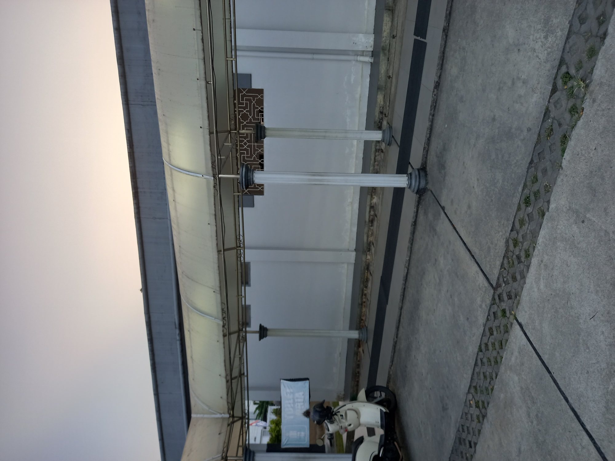 The image captures an outdoor scene, likely in Samarinda, Indonesia, featuring a covered walkway or shelter. The structure has a white, translucent roof supported by metal beams and white, decorative pillars. A beige scooter is parked on the concrete ground to the left of the frame. Behind the walkway, a plain white wall is visible, with a dark decorative panel featuring an interlocking geometric pattern mounted on it. A banner that reads "TOILET" and "WANITA" (woman in Indonesian) is displayed on the left side of the wall. The ground is paved with concrete, with a section of interlocking paving stones interspersed with grass running diagonally across the foreground. The sky above is a pale, soft gradient of light blue and pink, suggesting either dawn or dusk. The environment appears to be a public area, possibly near a facility that requires restrooms.