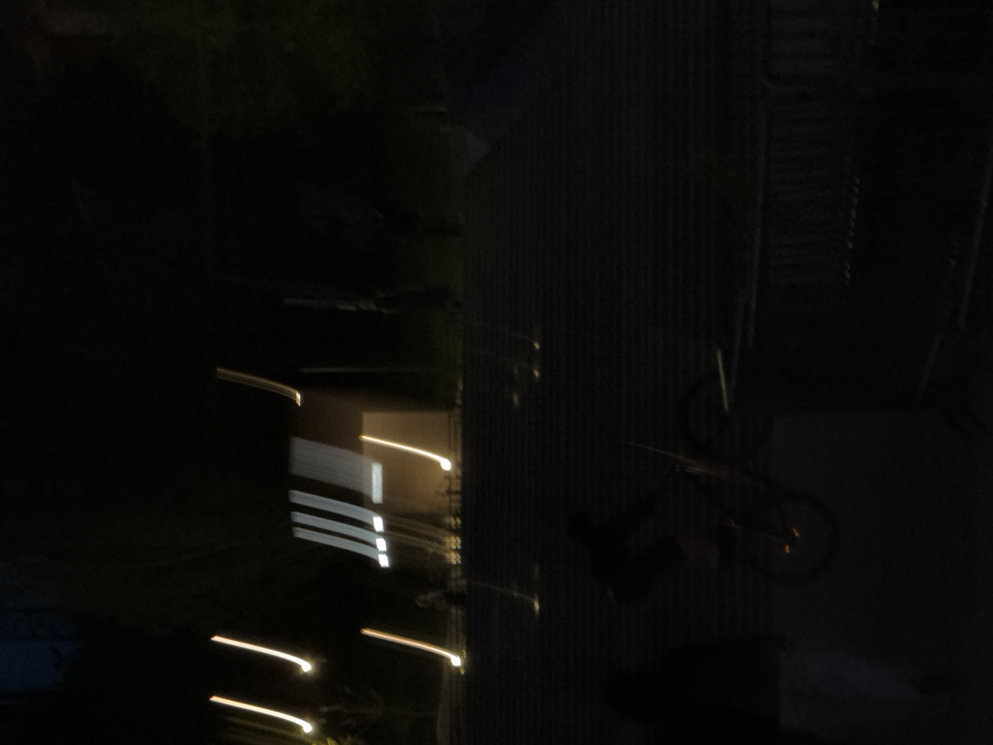 The image, likely taken at night in Izhevsk, Russia, shows a dimly lit urban scene. To the right, a corrugated metal structure is visible, possibly a wall or building facade. A bicycle leans against this structure. There is an indistinct dark figure standing near the bicycle, making it difficult to determine what action or interaction is occurring. To the left, there are streaks of light, suggesting passing cars or street lights. Overall, the scene is dark and blurry, and the information available is minimal.