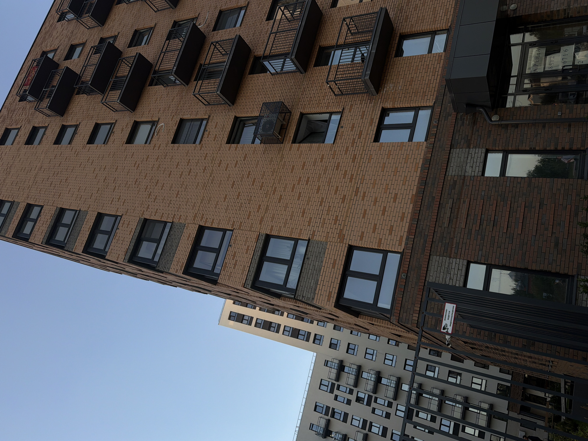 The image shows a low-angle view of two modern apartment buildings in Izhevsk, Russia. The taller building features light brown brickwork, rectangular windows, and projecting dark metal balconies. Adjacent to it is a building with darker brown brickwork. In the foreground, a dark fence partially obscures the view of a second, lighter-colored apartment building with similar window and balcony designs. The sky is a clear light blue. A surveillance sign is visible near the dark fence.