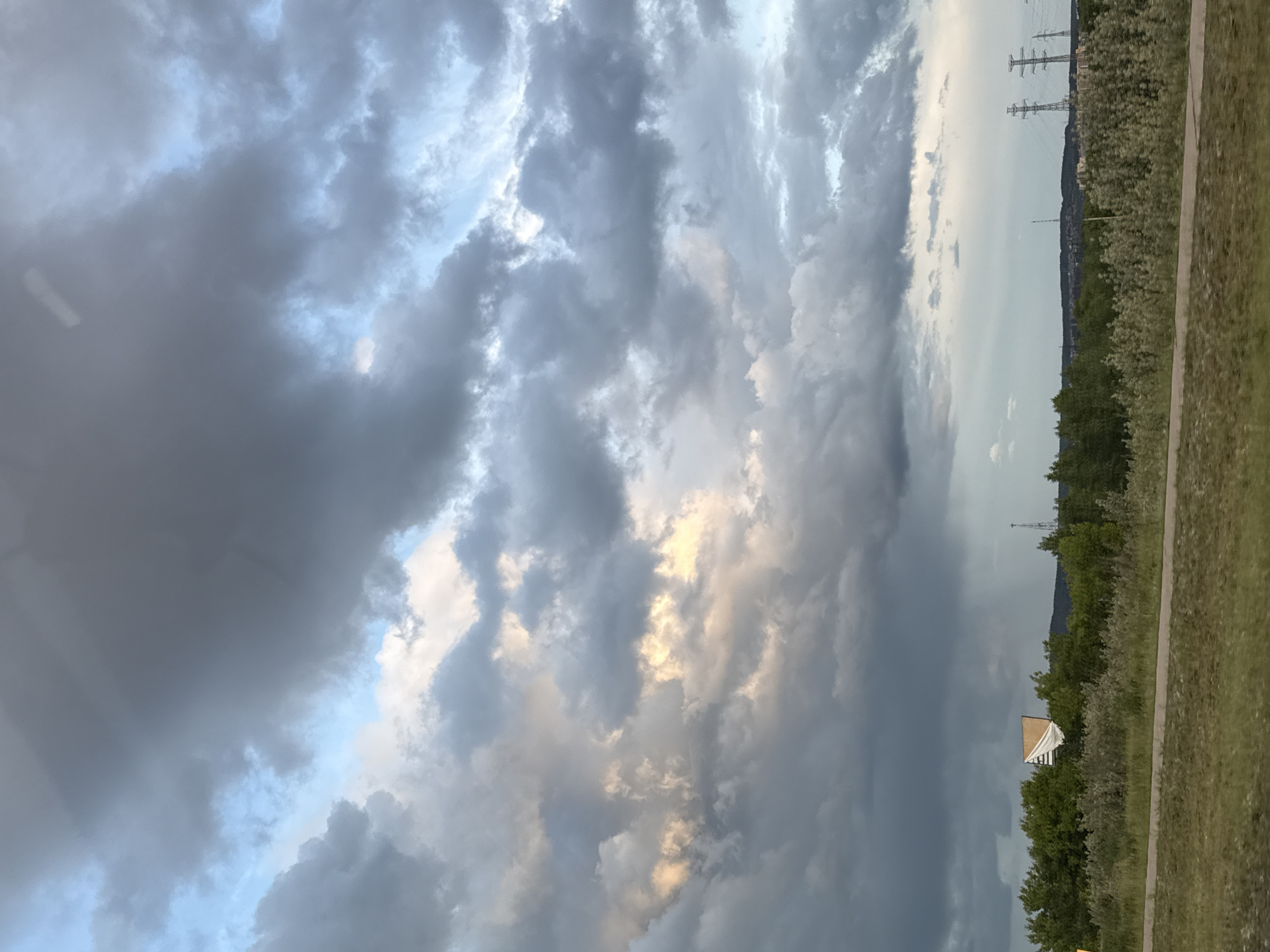 The image depicts a sky dominated by a dense cloud formation, with patches of light breaking through. In the background, a landscape of trees and vegetation stretches horizontally. Electrical power lines are visible on the horizon, suggesting an urban or semi-urban setting. A small building, likely a residential structure, is partially visible among the trees. The photograph was taken in the city of Chaikovsky, Russia.