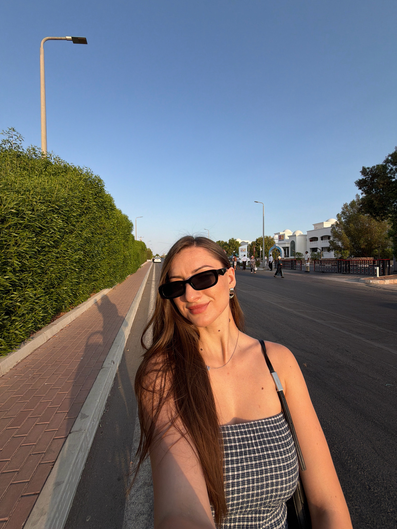 A young woman with long brown hair and black sunglasses takes a selfie while walking on a paved sidewalk next to a manicured green hedge. She is wearing a plaid top and a necklace. The sun is casting long shadows. In the background, there is a road with some cars and buildings, suggesting an urban or resort setting. The sky is clear and blue, indicating it is daytime. The location is Sharm El Sheikh, Egypt.