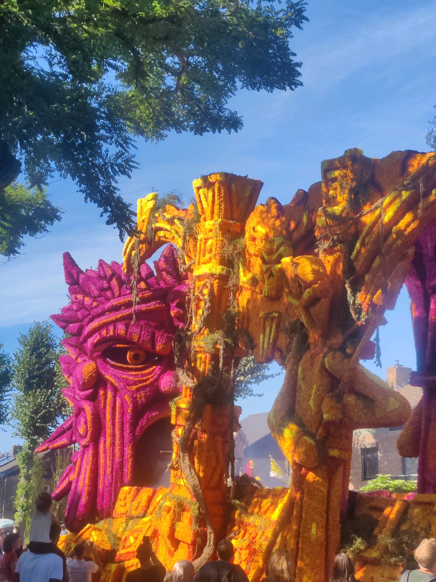 This is a vertical, full shot photograph taken during the daytime at the Flower Parade in Zundert, Netherlands. The main subject is a large, ornate float decorated with an abundance of colorful flowers. The float features a prominent, stylized face on the left side, predominantly adorned with vibrant pink and orange flowers, with a single large eye visible. To the right of the face, the float continues with an elaborate, abstract structure, primarily decorated in shades of orange and yellow flowers, with some green vines draped around it. The overall design suggests a mythical or fantastical theme.
The background includes a clear blue sky with scattered white clouds, and the green foliage of trees frames the top and left side of the image. In the lower part of the frame, a crowd of people are visible, suggesting they are spectators at the parade. Some individuals are looking towards the float, and one person is being carried on another's shoulders. The presence of buildings in the background, visible through gaps in the float's structure, indicates an urban or semi-urban setting within Zundert. The lighting suggests a sunny day, casting distinct shadows and highlighting the textures of the floral decorations.
