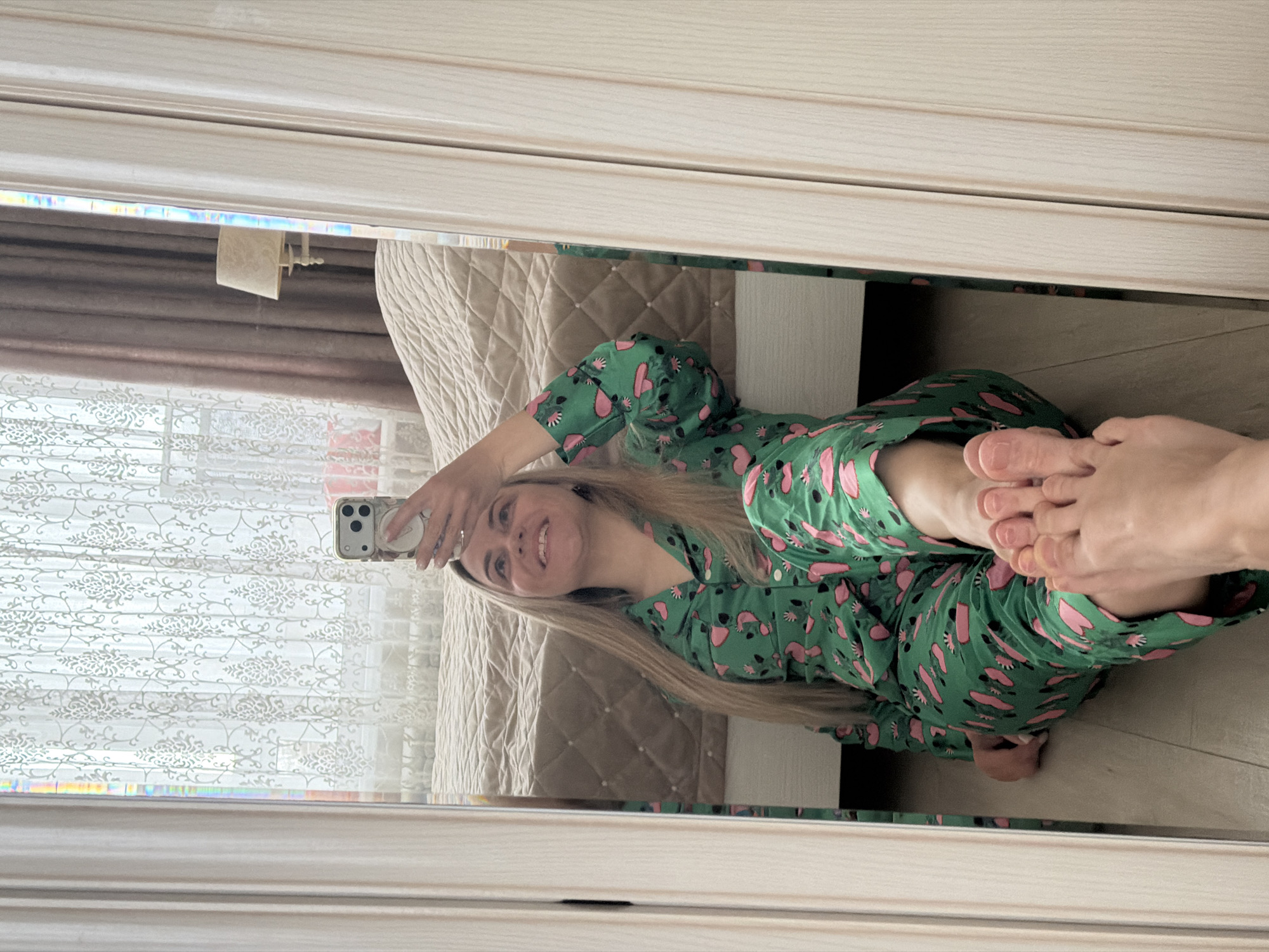 The media file displays a mirror selfie of a young woman taken indoors, likely in a bedroom or living space in Votkinsk, Russia. The original image is rotated 90 degrees clockwise, showing the woman lying or sitting on the floor.
The main subject is a young woman with long, straight blonde hair, smiling broadly at her phone. She is wearing a vibrant green pajama set adorned with a repeating pattern of pink heart-like shapes and other abstract motifs. Her left hand is holding a light-colored smartphone, which has multiple camera lenses and a white pop socket or grip accessory on its back. A ring is visible on her left ring finger. Her bare feet are also prominently featured in the lower right portion of the reflection.
The setting includes a large window to the left of the woman, covered by white sheer curtains with a patterned design, and partially visible darker drapes. Above the window, part of a blind mechanism or valance is visible. To her right, a bed with a plush, light-colored, quilted headboard and bedding is present. The floor where she is positioned appears to be a light-toned material, possibly wood or laminate. The entire scene is reflected in a large mirror, whose light-colored frame is visible at the top and bottom of the image.
The activity depicted is a casual mirror selfie, captured during what appears to be daytime, suggested by the natural light filtering through the window. The woman's wide smile conveys a relaxed and happy emotion. No visible text is present in the image.