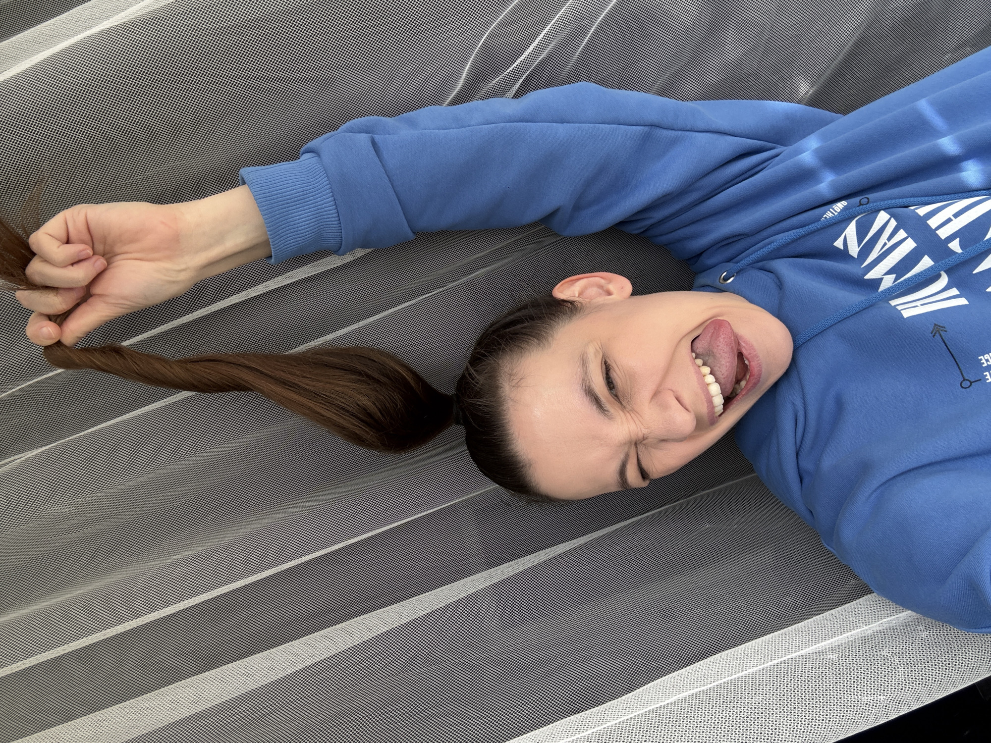 A young woman lies on a surface covered with a white mesh fabric, possibly a net or a screen. She wears a blue hooded sweatshirt with white writing. Her brown hair is pulled back into a ponytail, and she is holding the end of the ponytail in her right hand. She is making a silly face with her tongue sticking out and eyes squinted. The location is Chaikovsky, Russia.