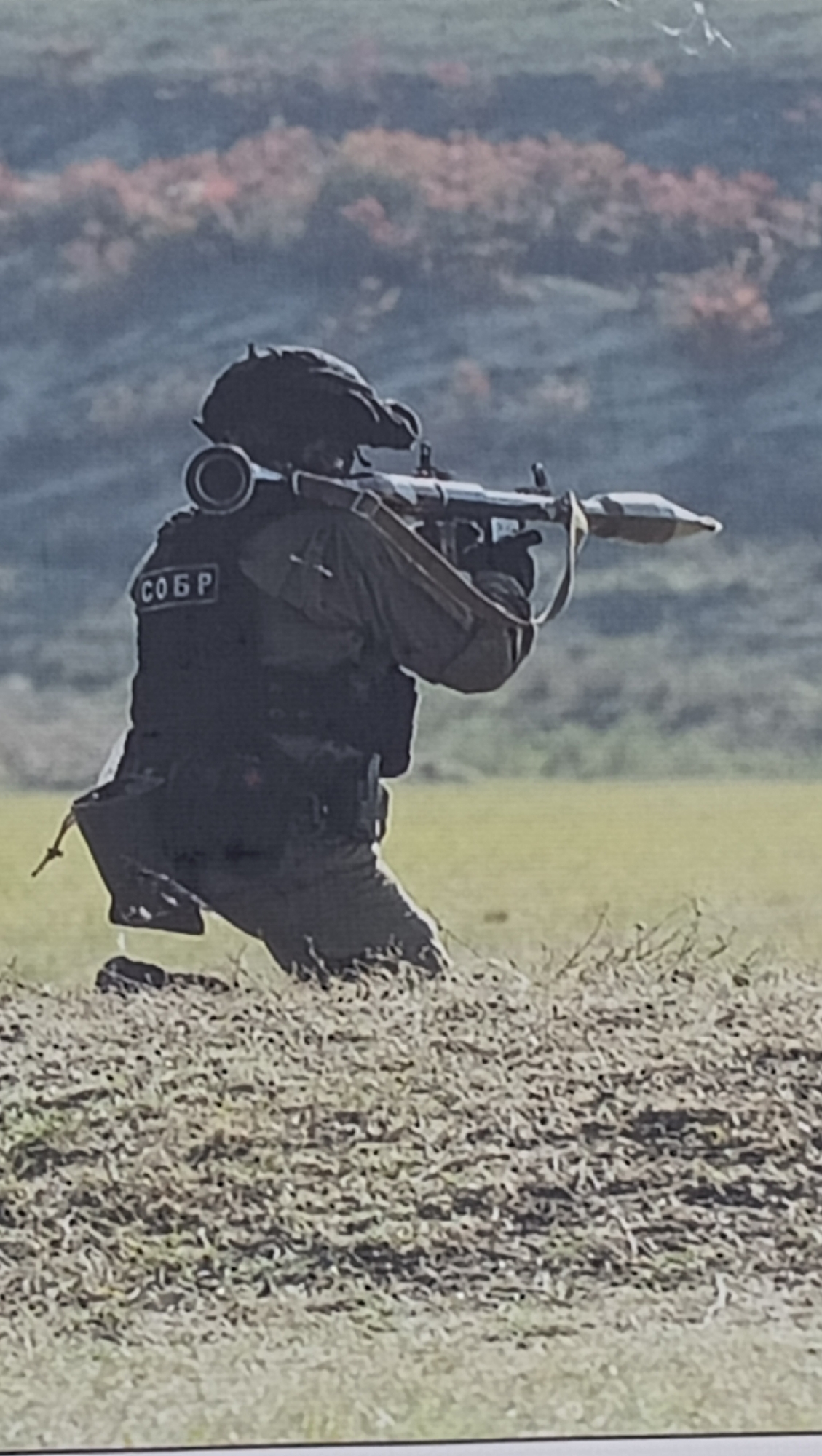 A person dressed in dark tactical gear, including a helmet, vest, and gloves, is depicted in a kneeling position outdoors, aiming a rocket-propelled grenade launcher (RPG) with a rocket loaded into the front. On the back of the person's vest, a patch with the Cyrillic text "СОБР" (SOBR, meaning Special Rapid Response Unit) is clearly visible.
The scene takes place during the daytime under natural light. The foreground and mid-ground are covered in dry, short grass. In the background, rolling hills or mountains are visible, featuring sparse vegetation with some reddish-brown tones, suggesting an autumn landscape. A few small, white specks, possibly birds, are high in the distant background sky.
The activity shows the individual in a posture of readiness, aiming the weapon, indicative of a military exercise, training, or operational deployment. Given the provided context, the image likely originates from Tiraspol, Moldova, and features a member of a local or regional Special Rapid Response Unit. The overall impression is one of tactical focus and preparedness.