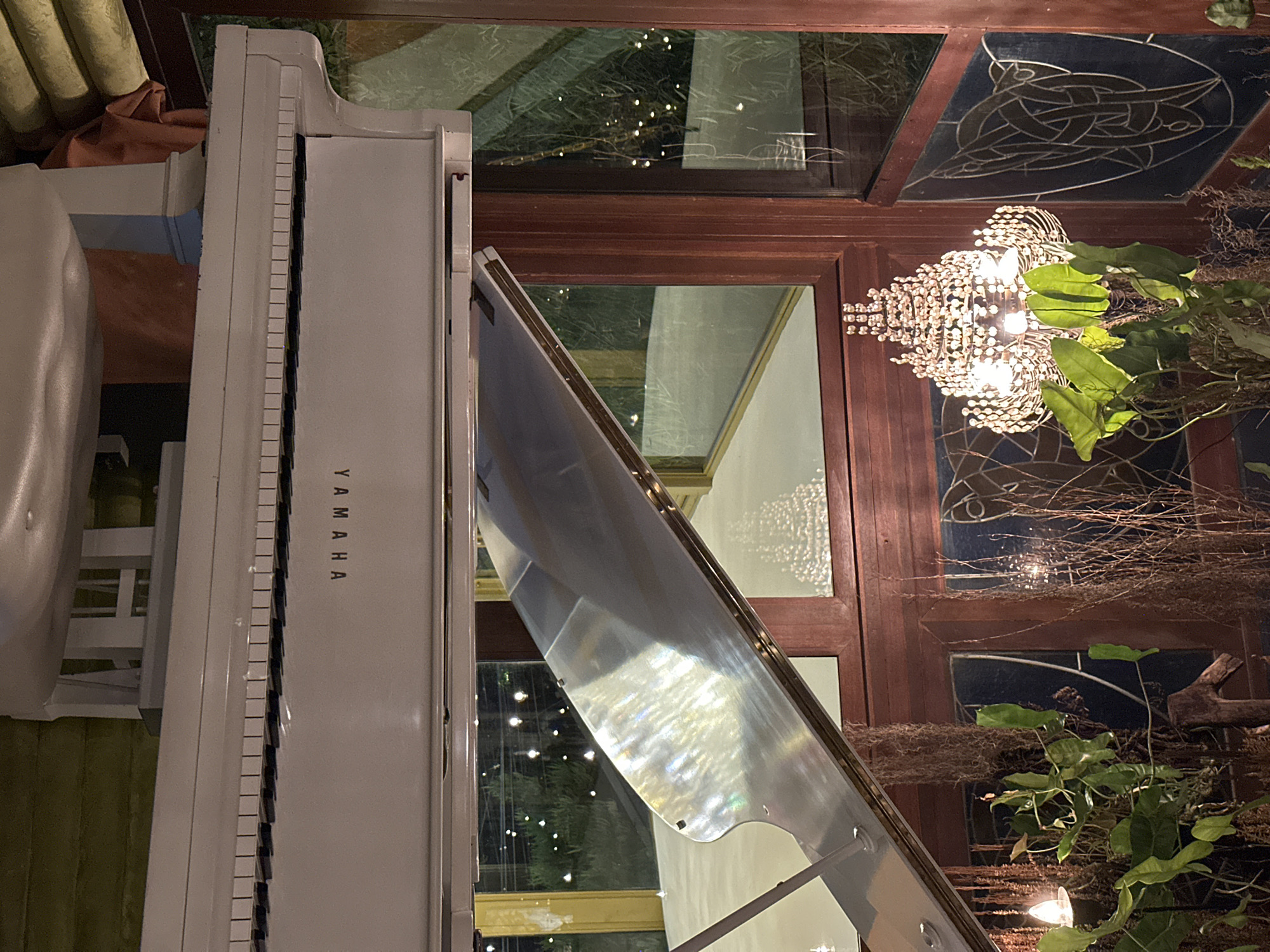 A white Yamaha piano is positioned near a window in Ubud District, Indonesia. The window has a brown wooden frame with decorative stained glass on the top and side sections. A chandelier hangs in front of the window, partially obscured by green plants. Outside the window, trees with small lights are visible.