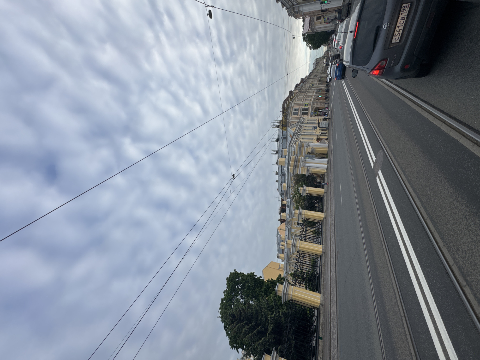 The image captures an overcast daytime street scene in Saint Petersburg, Russia, viewed from a slightly elevated perspective. The street features a wide asphalt road with dual tram tracks running down the center, flanked by a lane marked with double white lines.
On the right side of the road, a queue of cars is visible. The most prominent vehicle is the rear of a dark grey Nissan Qashqai, displaying the Russian license plate "C541CB 198", with "198" indicating its registration in Saint Petersburg or the Leningrad Oblast. Further ahead, a blue car and other vehicles can be seen.
To the left of the road, a long row of multi-story buildings stretches into the distance. These structures are predominantly light yellow or cream-colored, characterized by classical architectural elements such as columns and ornate black metal fences with yellow posts. A large, dense green tree stands in the foreground, partially obscuring the buildings behind it.
The sky is heavily overcast, filled with a pattern of rippled, altocumulus-like clouds. Numerous overhead wires, likely for tram power or utilities, crisscross the expanse of the sky. The overall impression is that of a bustling urban environment under a cloudy sky.