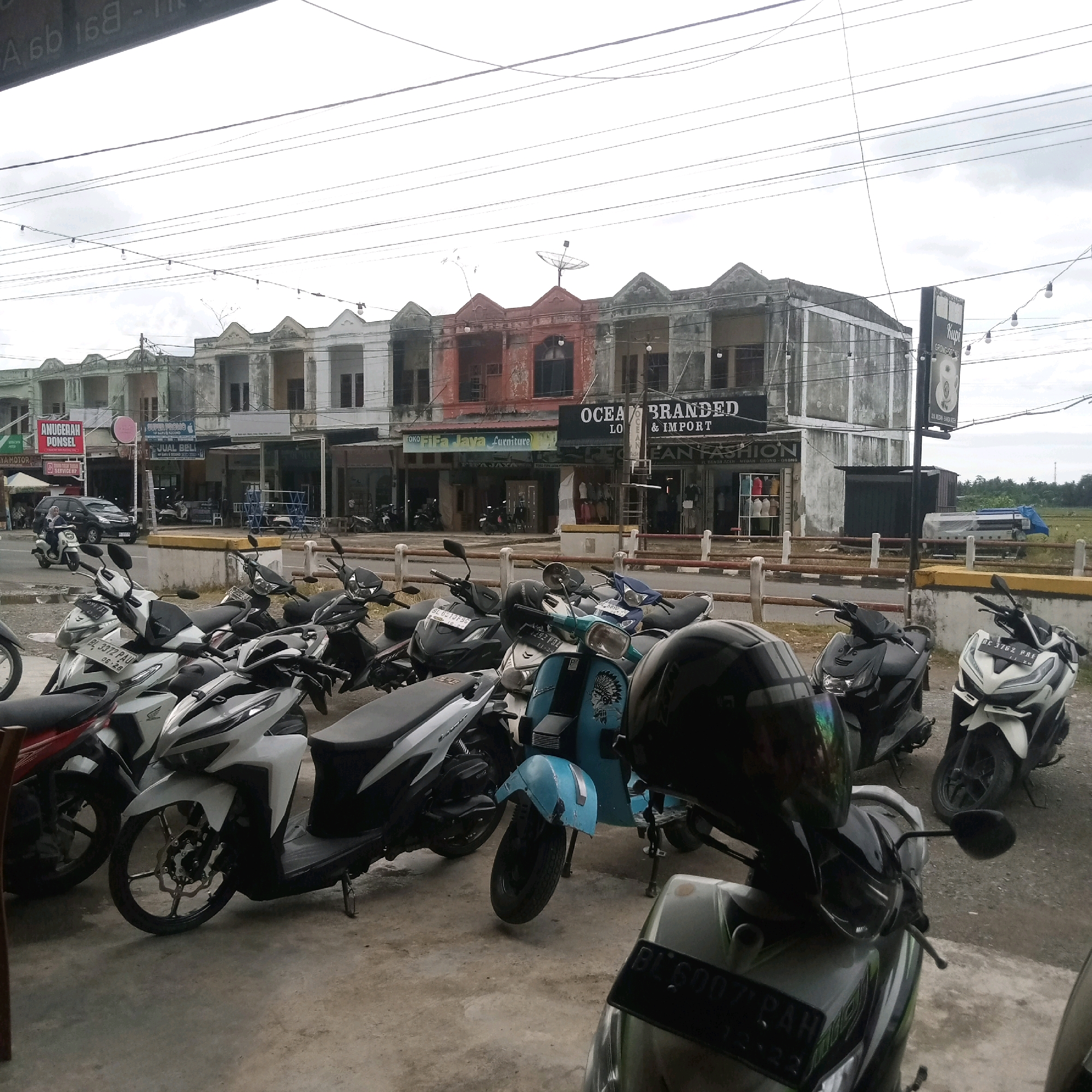 The image depicts a street scene in Grong-Grong, Indonesia, on a cloudy day. In the foreground, a row of parked motor scooters and motorcycles occupies a paved area. These vehicles are varied in color and style, with some displaying license plates. Behind the vehicles, a row of modest, two-story buildings lines the street. These buildings have a weathered appearance, with some showing signs of fading paint and older architectural styles. Signs are visible above the storefronts, indicating businesses such as "Fifa Jaya Furniture" and "Ocean Branded," along with others that are less legible. A black car and a person riding a white scooter are visible on the road in the middle ground. Power lines are strung across the sky above the buildings. In the background, a vast, flat field stretches to the horizon, with a few scattered trees and a large printer or industrial machine covered by a blue tarp. The overall atmosphere is one of a typical commercial street in a provincial town.