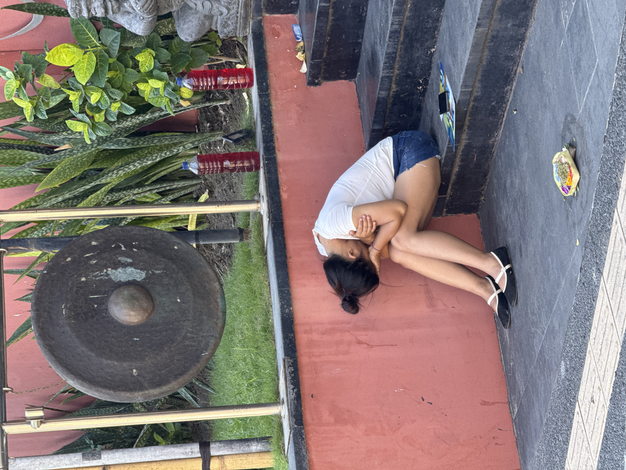 A young woman wearing a white t-shirt and denim shorts sits slumped on dark grey stone steps, her face buried in her arms. Her dark hair is pulled back into a bun. Black ballet flats with white straps are on her feet. To her right, a stack of dark grey steps ascends. On the steps, a thin booklet or flyer is open, topped with a black mobile phone. Further up the steps, two red plastic bottles containing a red liquid are visible, along with a small pile of yellow and blue wrappers. Behind the woman, a textured, pale pink wall rises, with a large, dark metal gong hanging on a frame to her left. Green grass is visible behind the gong, and a dense planting of snake plants and a smaller leafy plant with bright green leaves are situated behind the bottles and to the right of the gong, against the pink wall. A small, woven basket containing colorful offerings of flowers and other items rests on the ground in front of the steps. A stone sculpture of a seated figure is partially visible on the far right, behind the plants and bottles. The scene appears to be in Denpasar, Indonesia, likely near a temple or public space given the offerings and gong.
