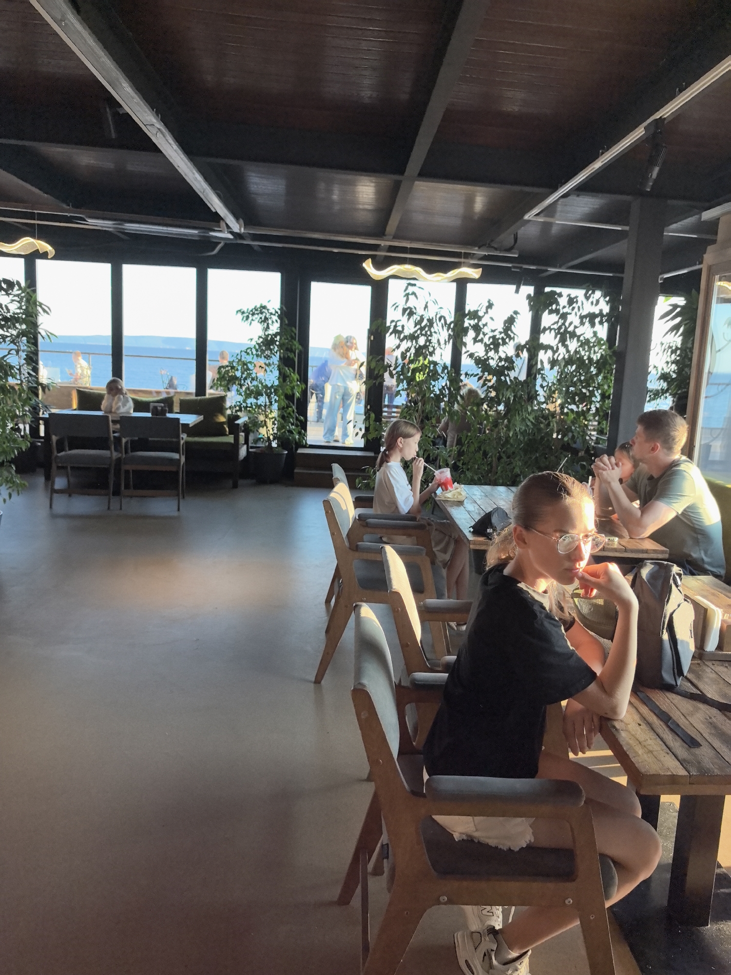 In a sunlit, contemporary restaurant with a view of a body of water, likely the Volga River given the Tolyatti location context, several people are enjoying a meal. The setting features large windows that allow ample natural light to flood the space, illuminating the wooden tables and chairs. Lush green plants are strategically placed throughout the restaurant, adding a touch of nature to the indoor environment.
In the foreground, a young woman with blonde hair and glasses is seated at a table, looking to her right with a pensive expression. She is wearing a black t-shirt and white shorts. Behind her, a man and a young girl are seated at another table, appearing to be engaged in conversation. The girl is wearing a green t-shirt and has her hands clasped together.
Further back, near the windows, a young girl with blonde hair pulled back in a ponytail is drinking from a red beverage with a straw. She is wearing a light-colored t-shirt. In the background, through the large windows, several people are visible on an outdoor terrace, enjoying the view of the water and the sky. The sun is casting long shadows, indicating it is late afternoon or early evening. The overall atmosphere seems relaxed and pleasant. The scene captures a casual dining experience in a scenic location.
