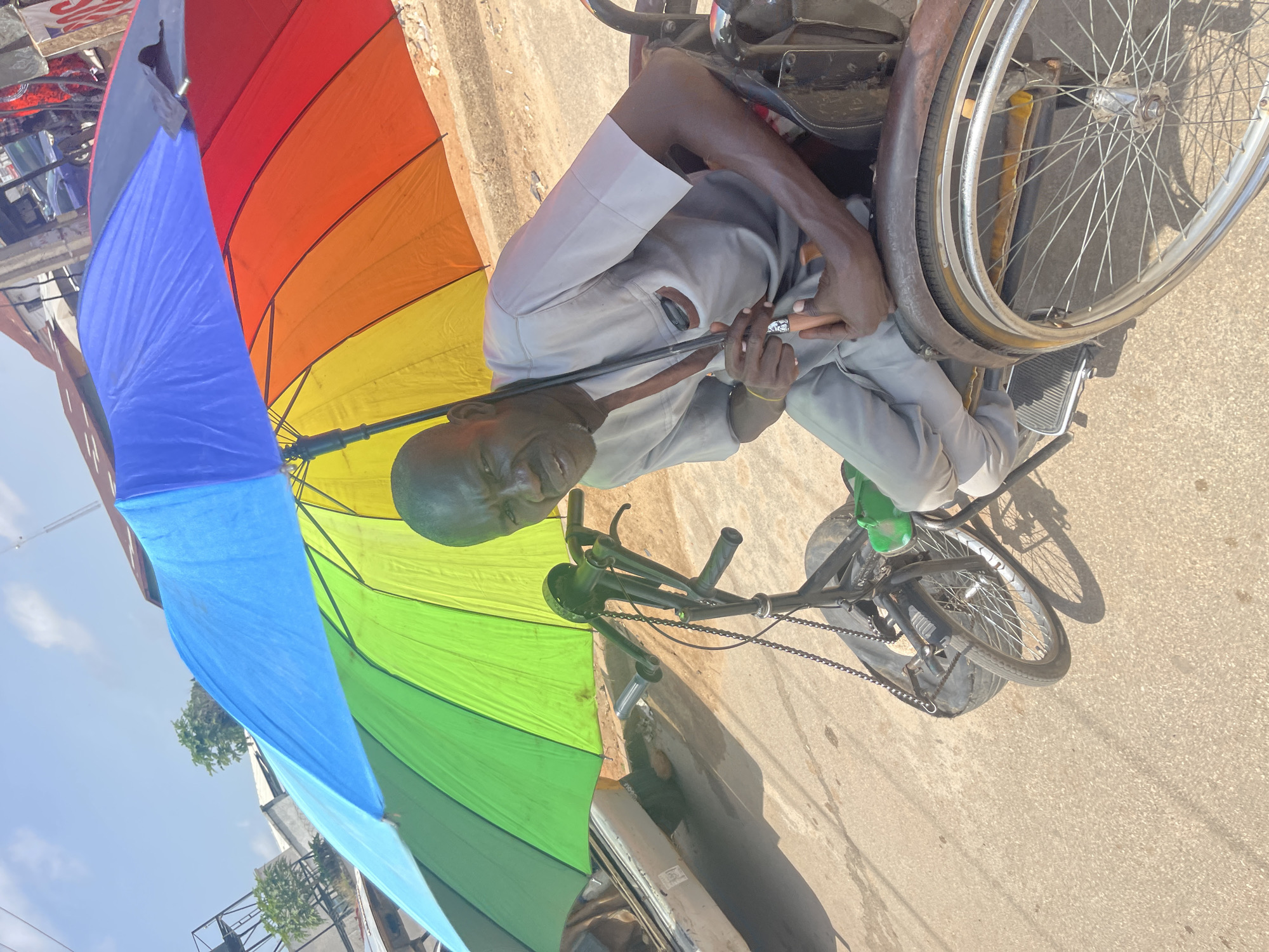 An elderly Nigerian man, seated in a wheelchair under a large, multi-colored umbrella, poses for a photo on a dusty, sun-drenched street in Jos, Nigeria. The man, with dark skin and a stern expression, wears a light gray tunic and trousers. He holds a closed black umbrella with a brown handle in his right hand. The wheelchair, appearing to be modified with bicycle parts, has large spoked wheels and a smaller front wheel assembly. Behind the man, a pale car and other vehicles are visible, suggesting a commercial or residential street. The background includes buildings and a clear blue sky, indicating a daytime setting.