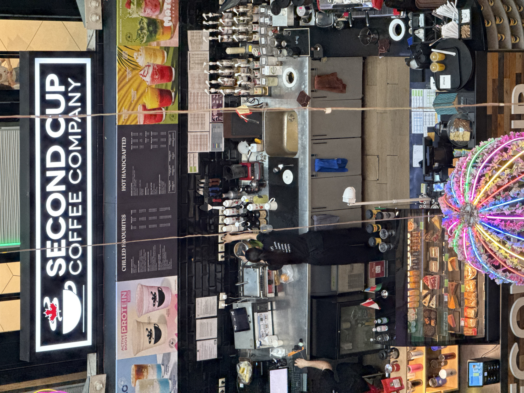 An overhead shot captures the interior of a Second Cup coffee shop, likely located in Baku, Azerbaijan, with a prominent illuminated sign featuring the brand's logo and name. The counter area is the primary focus, where a female barista, wearing a black uniform with "H! Lattes" text on the back, is actively engaged in preparing drinks. She is facing away from the camera, her hands occupied with tasks behind the counter.
Various coffee-making equipment and supplies are visible, including espresso machines, blenders, sinks, a variety of syrups in bottles, and cups. On the wall behind the counter, menu boards are displayed, listing "CHILLED FAVOURITES" and "HOT HANDCRAFTED" beverages, alongside promotional images of drinks.
To the left of the counter, a display fridge showcases an assortment of pastries and cakes. Further to the left, a shelf holds packaged coffee products and other retail items. In the foreground, a brightly lit, colorful neon light fixture, resembling a sphere adorned with flowers, adds a decorative element to the scene. Two small flags, one white and red and another green, white, and red, are positioned on the counter.
The overall environment appears to be a clean and organized retail space, typical of a coffee chain. The lighting suggests an indoor setting, possibly during daytime hours due to the ambient light filtering from above. The presence of flags and the context of coffee shops being common in Baku suggest this location is indeed in the capital of Azerbaijan.