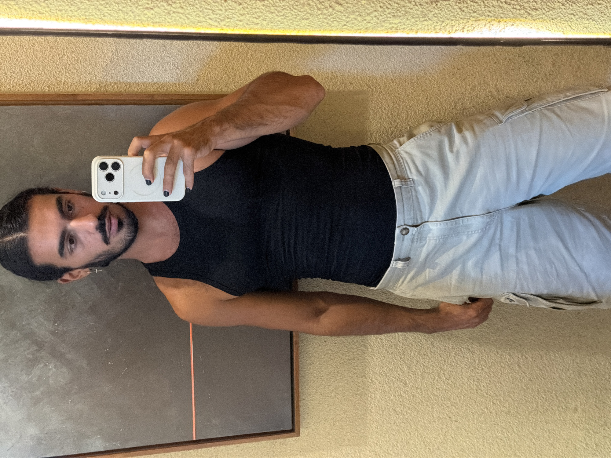 A young man with dark hair pulled back and a beard takes a selfie in a room. He is wearing a black tank top and light-colored cargo shorts. He is holding a white smartphone in his right hand. A framed abstract art piece with a red stripe hangs on the wall behind him. The wall has a textured finish. A bright, yellow-lit vertical fixture is visible to the right of the man.