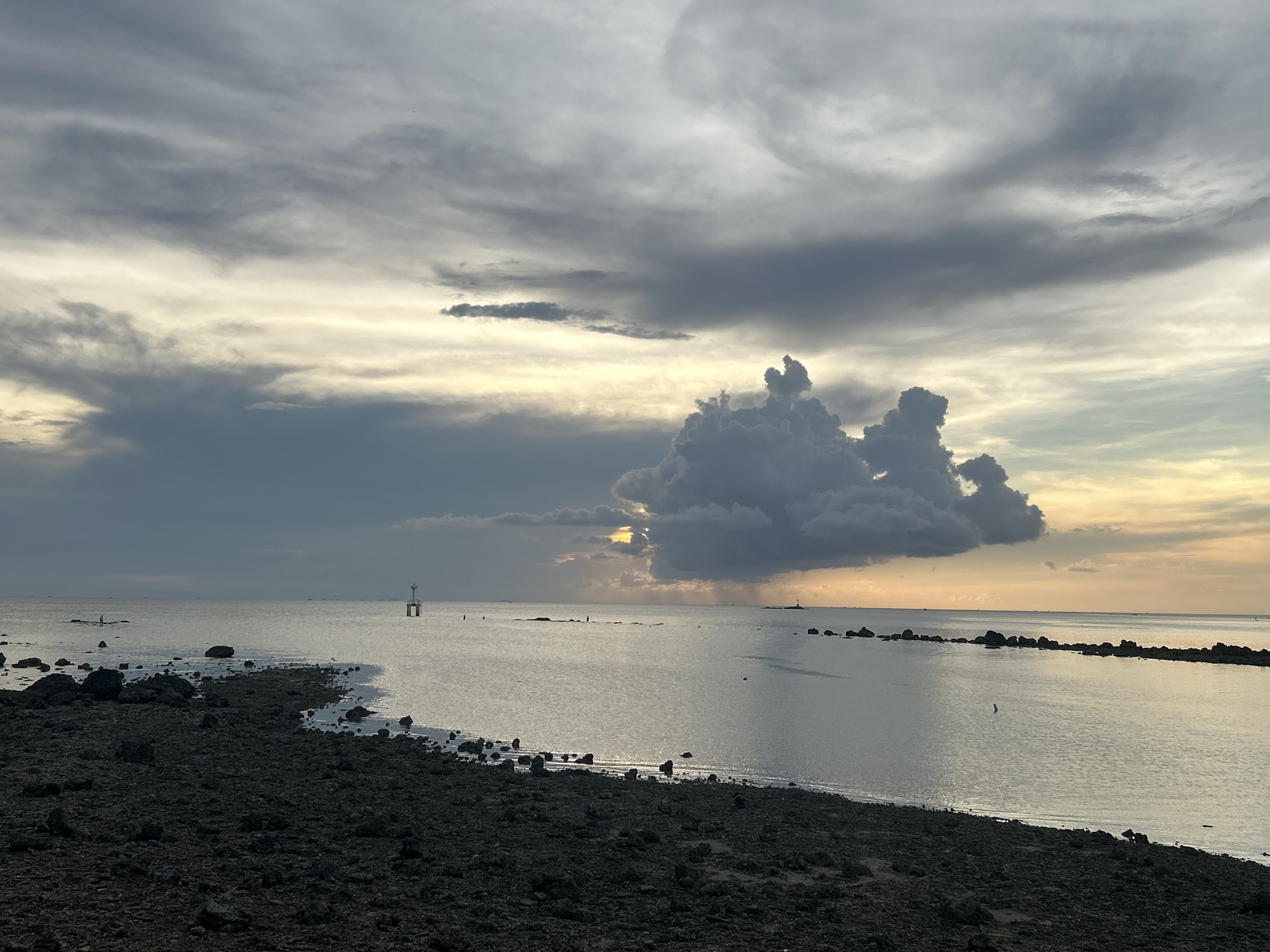 The image shows a coastal scene near Baan Sri Thanu, Thailand. The foreground is a dark, rocky beach transitioning to a calm sea. In the water, there's a small navigational marker resembling a tower. Clouds fill the sky, with a particularly prominent, large cloud formation appearing near the center of the frame, obscuring the sun partially. The overall lighting suggests either sunrise or sunset. The water reflects the sky's colors, creating a silvery effect. A few scattered rocks are visible in the water, and a line of rocks extends to the right forming a pier. There appears to be a person standing on some rocks in the distance towards the left.